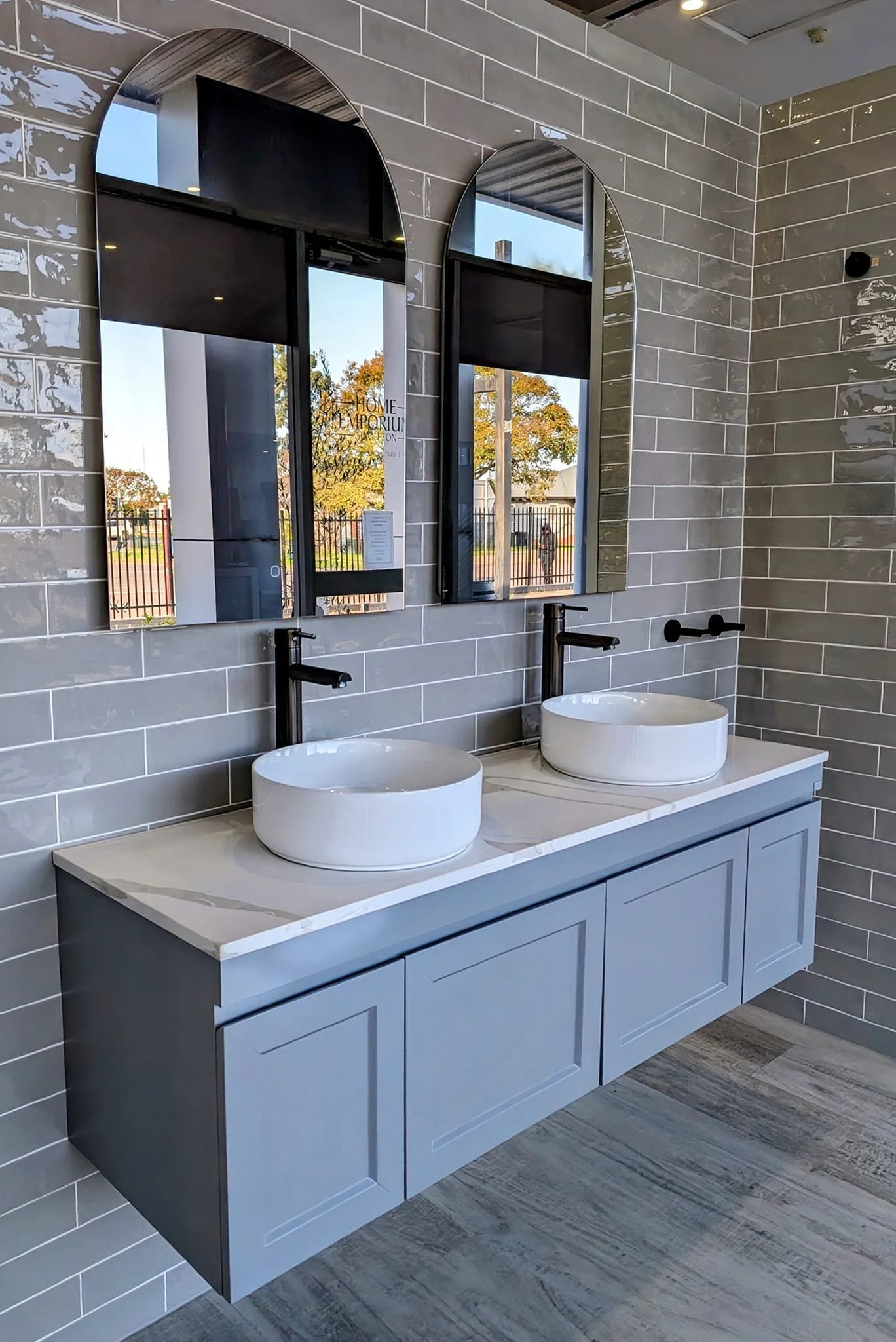 Modern bathroom vanity with two white vessel sinks, black wall-mounted faucets, two large mirrors with arched tops, gray cabinetry, gray tiled walls, and wood-look flooring.