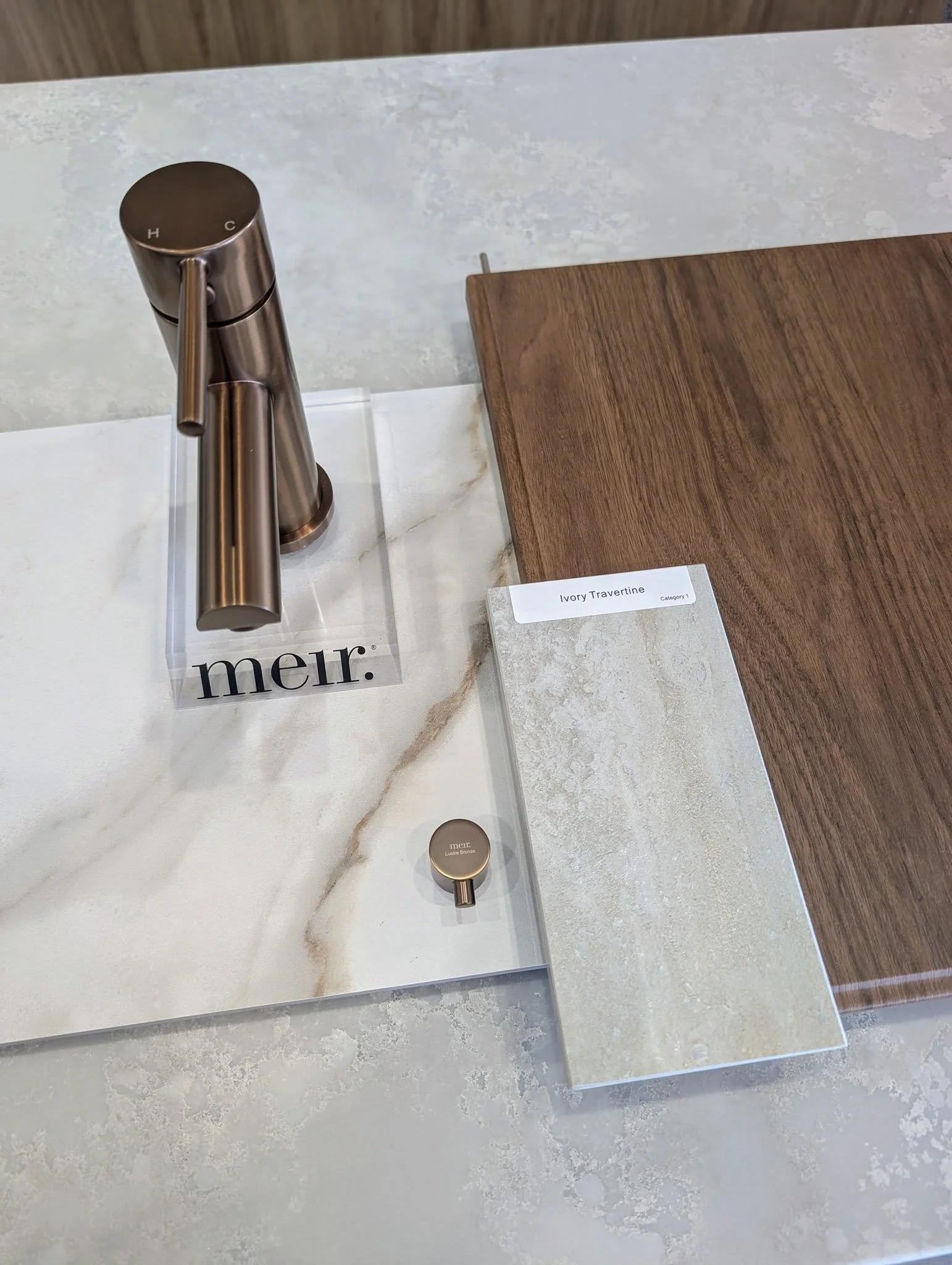 Close-up of kitchen countertop with a modern metallic faucet, a wood cutting board, and a small round metal object labeled "meir." on display in a store.
