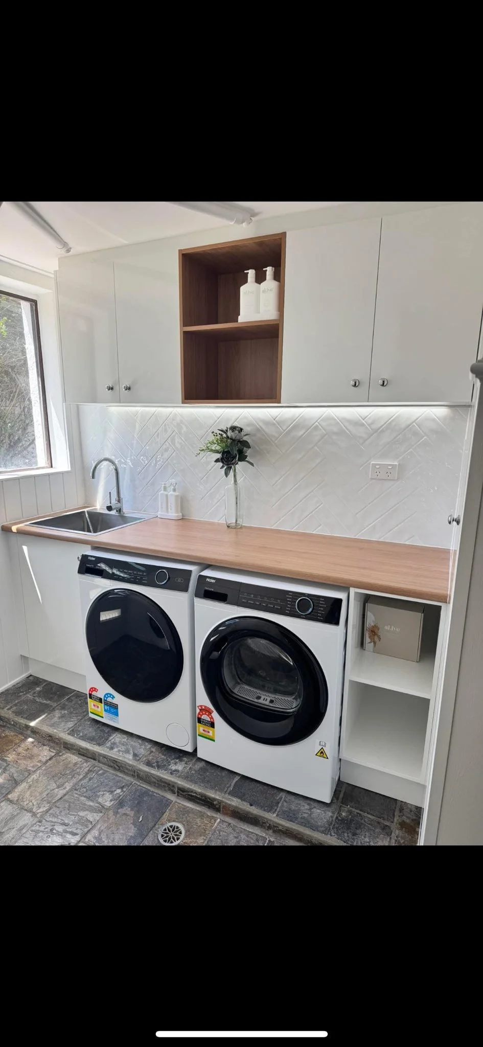 A laundry room with a wooden countertop, white cabinets, a stainless steel sink, a vase with flowers, and a washing machine and dryer.
