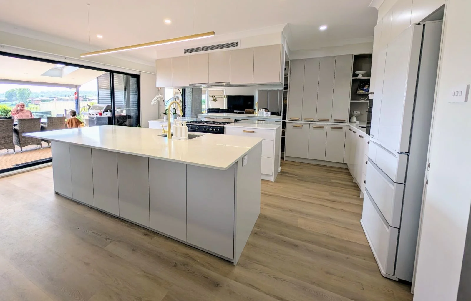Modern kitchen with white cabinets, a large island with a white countertop, and hardwood floors. A sliding glass door leads to an outdoor patio with people and outdoor furniture. The kitchen includes a built-in oven, a gas stove, a refrigerator, and open shelving.