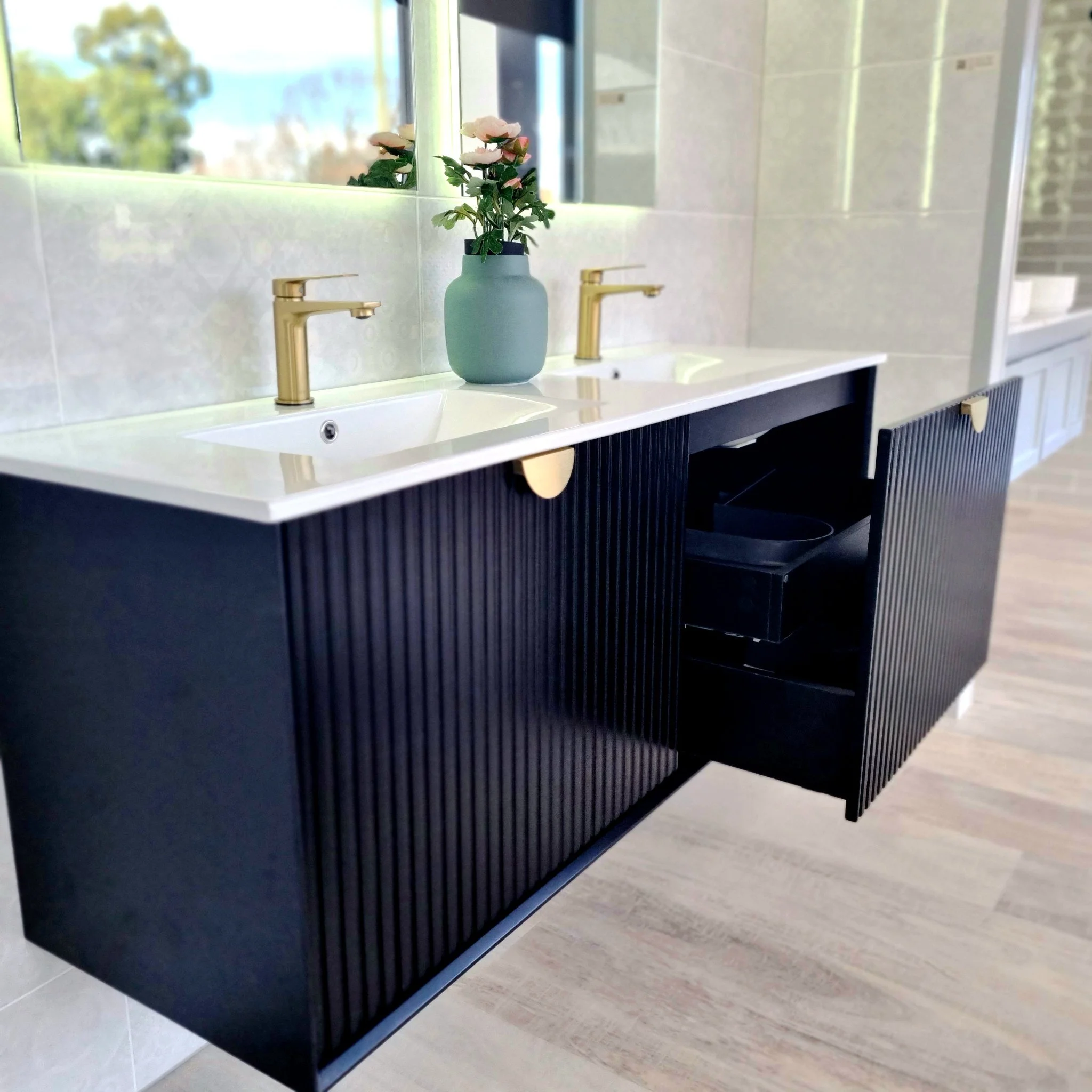 Bathroom vanity with two sinks, gold faucets, mirrored backsplash, and a blue vase with pink flowers in between the sinks.