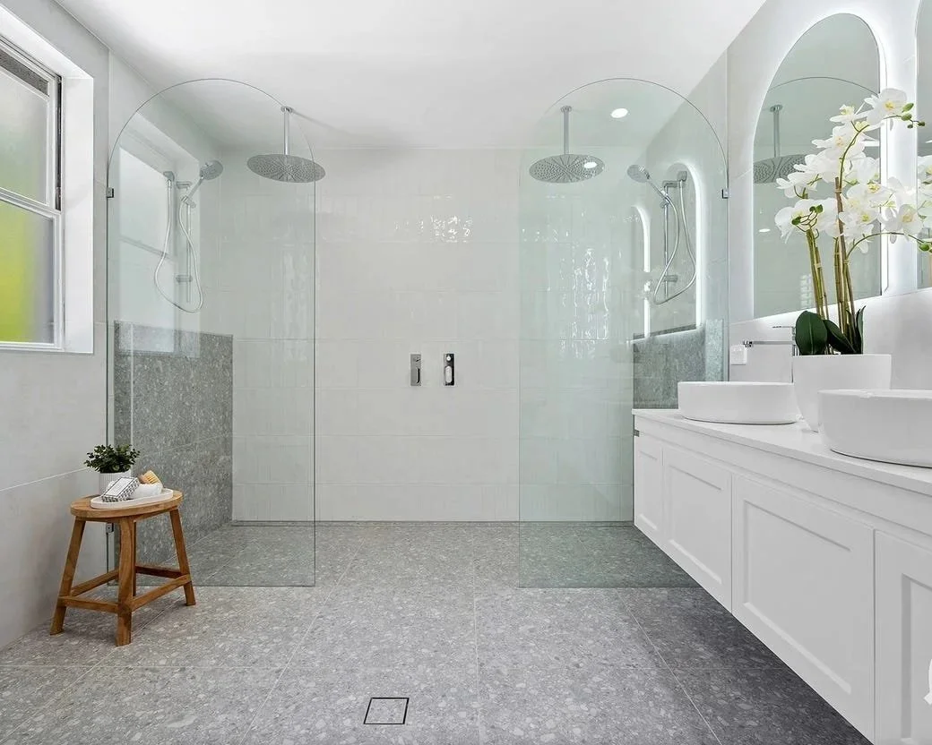 Modern bathroom with a walk-in glass shower, white cabinetry with two vessel sinks, a round mirror, and a potted orchid on the countertop. There is a small wooden stool in the corner with a plant and a book. A window provides natural light.