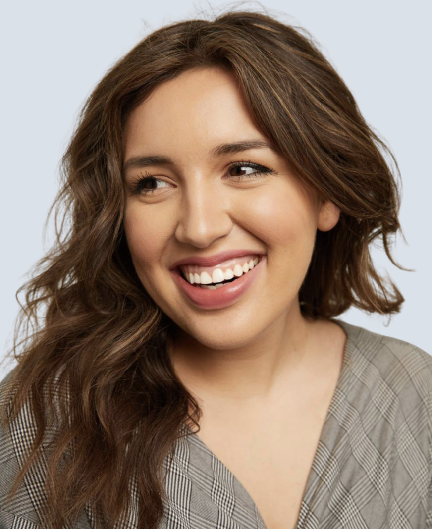 A young woman with wavy brown hair smiling and looking slightly to her right, wearing a gray checkered top.