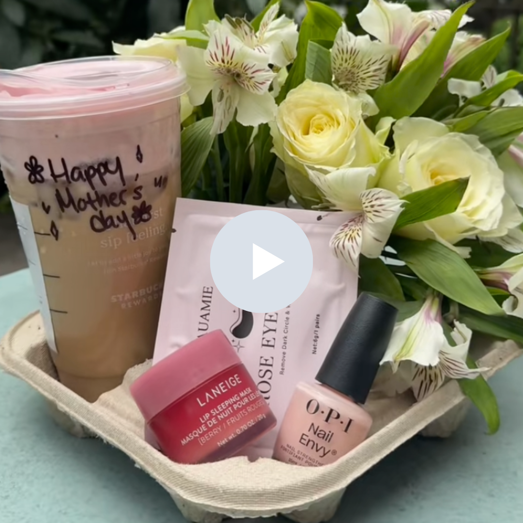 A Mother's Day gift basket with a coffee tumbler, skincare products, and a bouquet of white and pink flowers.