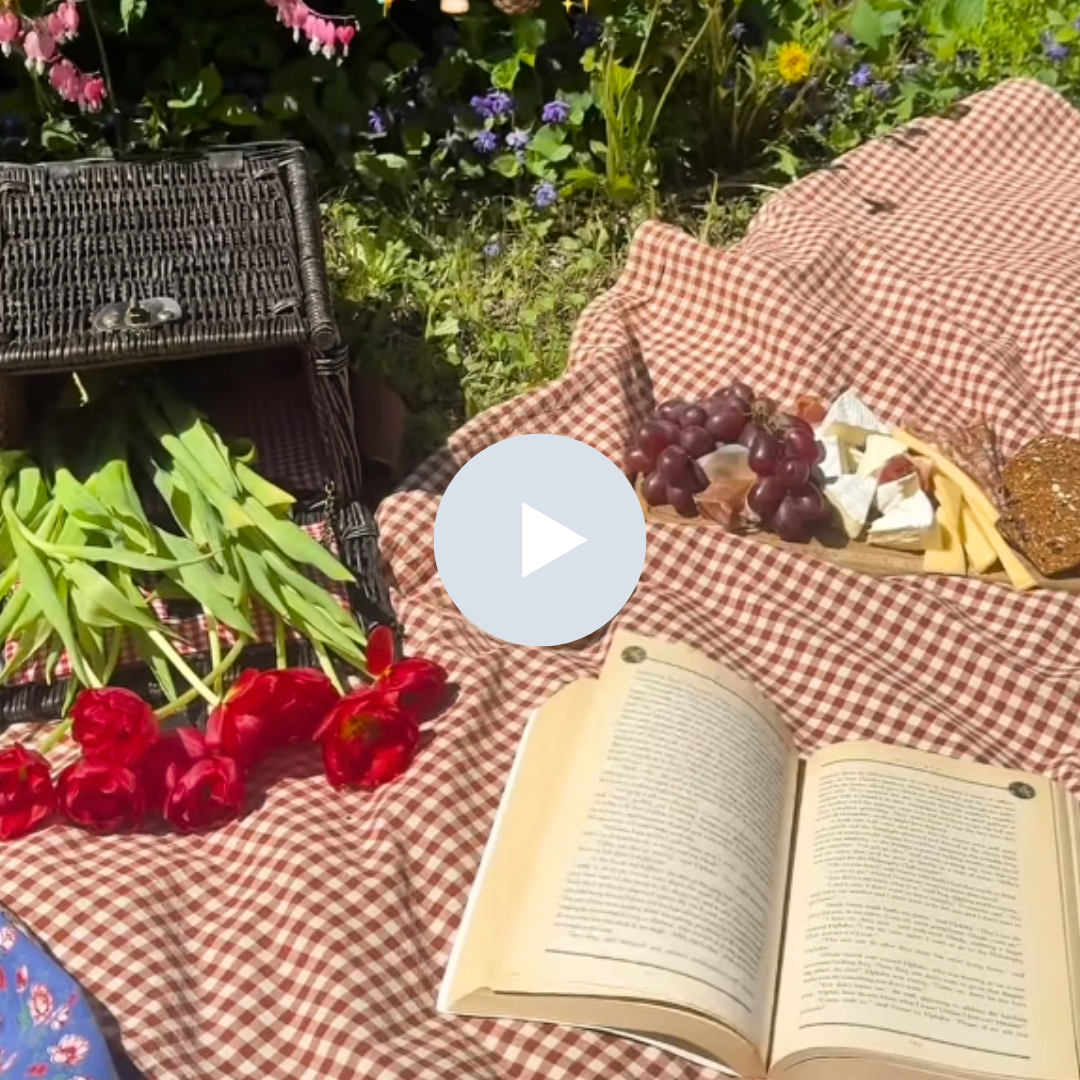 A picnic setup on a checkered tablecloth with red flowers, green leafy vegetables, an open book, a bunch of grapes, cheese, bread, and a basket of assorted items, surrounded by colorful flowers and greenery.