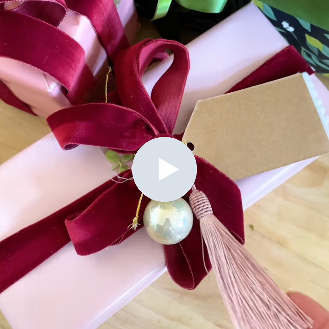 Close-up of a gift box wrapped in pink paper with a burgundy velvet ribbon bow, decorated with a large pearl, a pink tassel, and a blank brown gift tag.