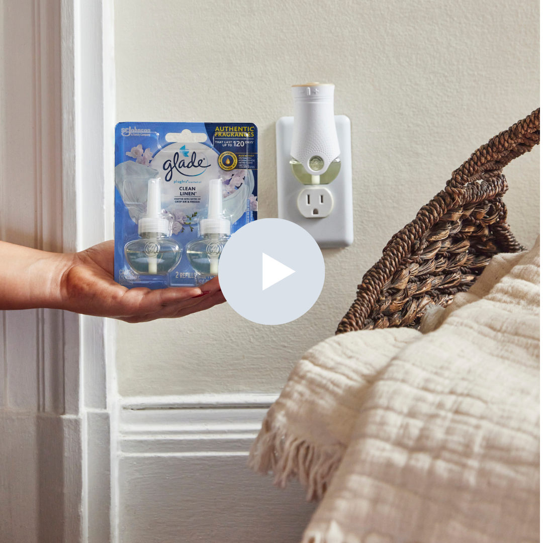 Person holding a package of Glade plug-in refill air fresheners in front of an electrical outlet with a plug-in air freshener and a wicker basket with a blanket nearby.