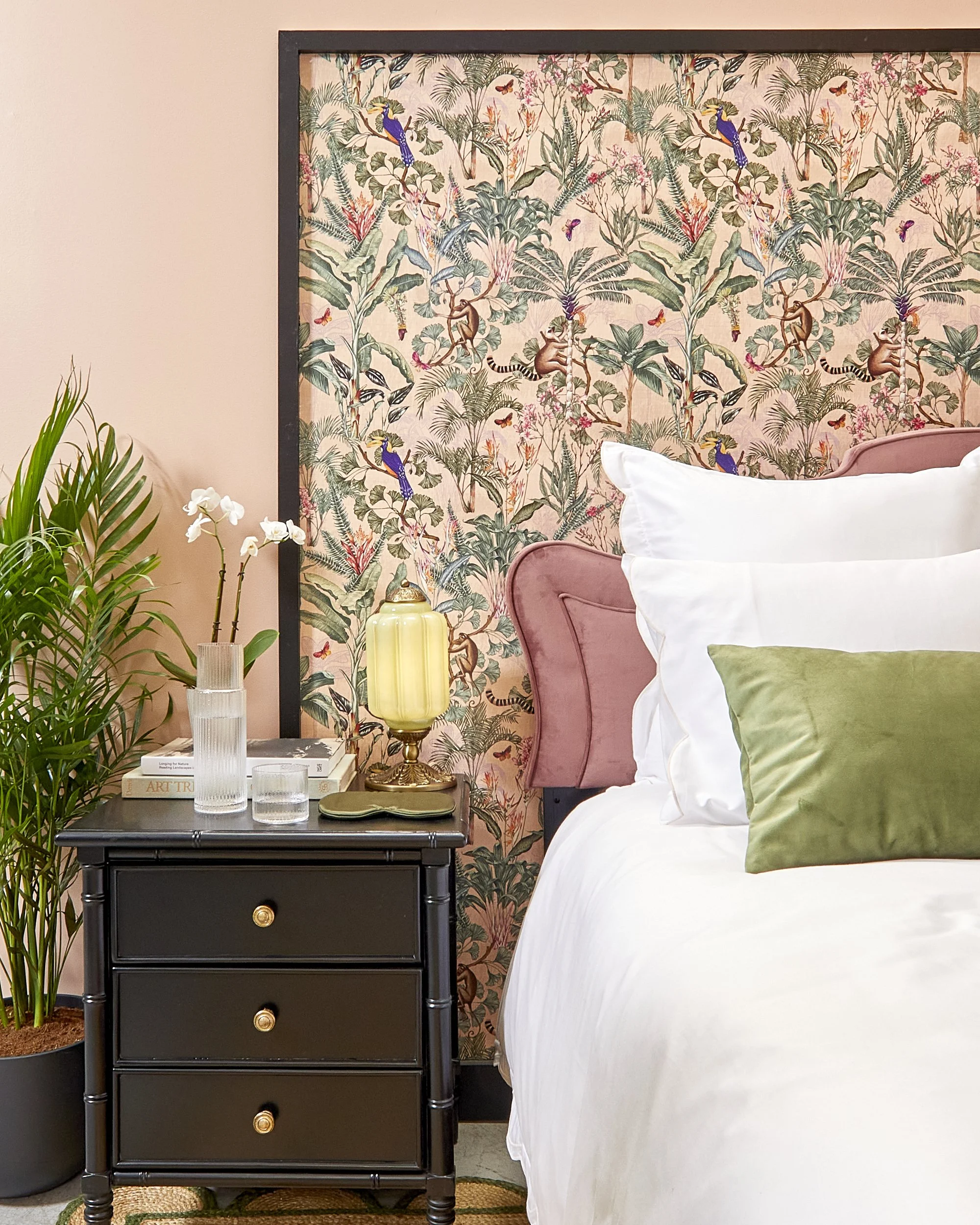 A bedroom featuring a black nightstand with three drawers, a yellow vintage lamp with a brass base, two glass of water, and a stack of books on top. Next to the nightstand is a potted green plant with white orchids. The background includes a patterned tropical wallpaper with birds, monkeys, and butterflies, a pink velvet headboard, white pillows, and a green pillow on a white bedspread.