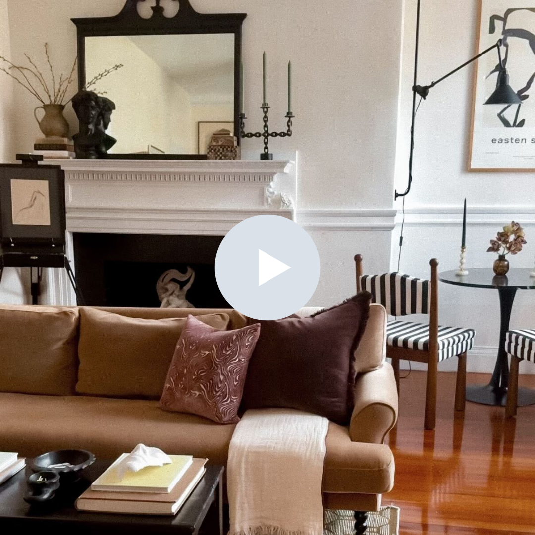Living room with a beige sofa, decorative pillows, a fireplace mantel with a mirror, books, a bust sculpture, and a candle holder. There is a small round table with a vase, a striped chair, and wall art with a wall-mounted lamp.