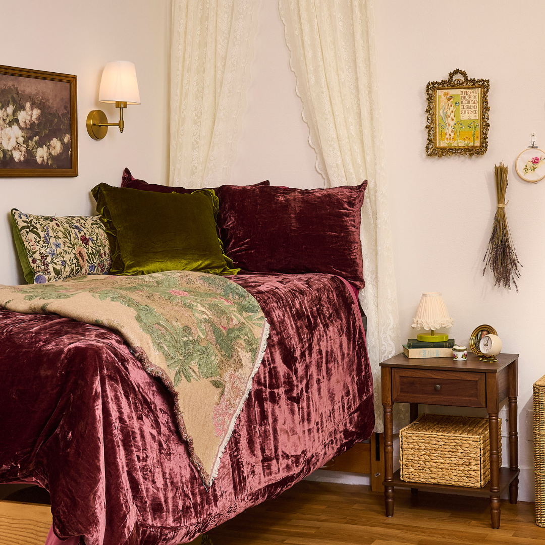 A vintage bedroom corner with a bed draped in a velvet pink bedspread and pillows in green velvet, burgundy, and floral patterns. A wooden nightstand holds a small lamp, books, and a clock. The wall features framed artwork, a wall sconce, and decorative hanging items including dried lavender and embroidery hoops.