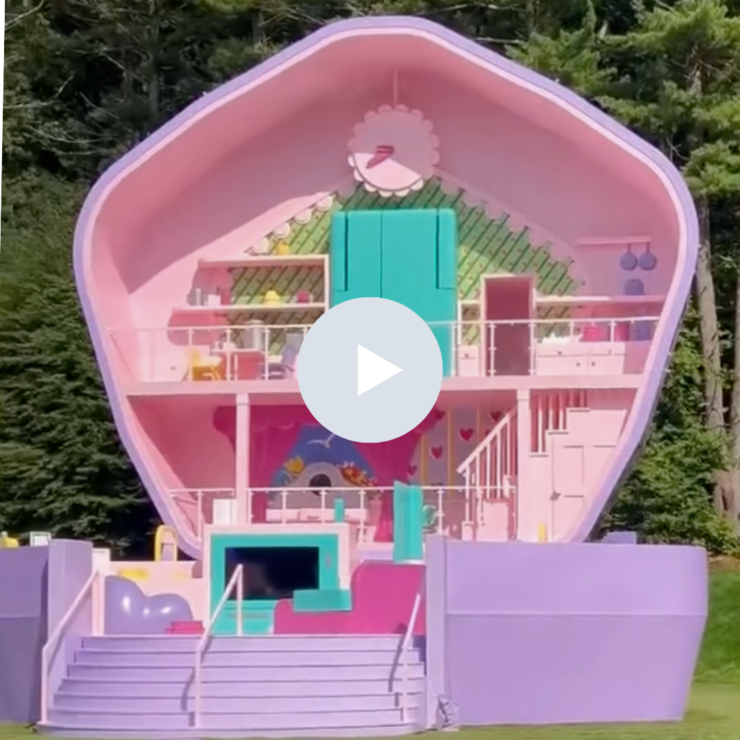 A pink dollhouse shaped like a seashell with multiple rooms and staircases, decorated with pink, turquoise, and pastel colors, set outdoors with trees in the background.