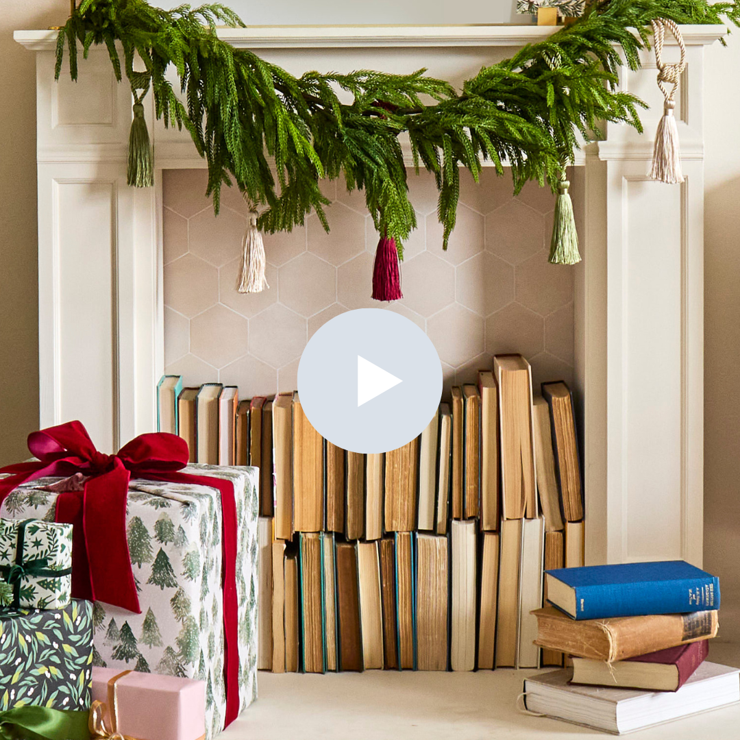 A decorated fireplace mantel with a green garland and tassels, holiday presents with ribbons and bows, and a stack of books placed in front of the fire