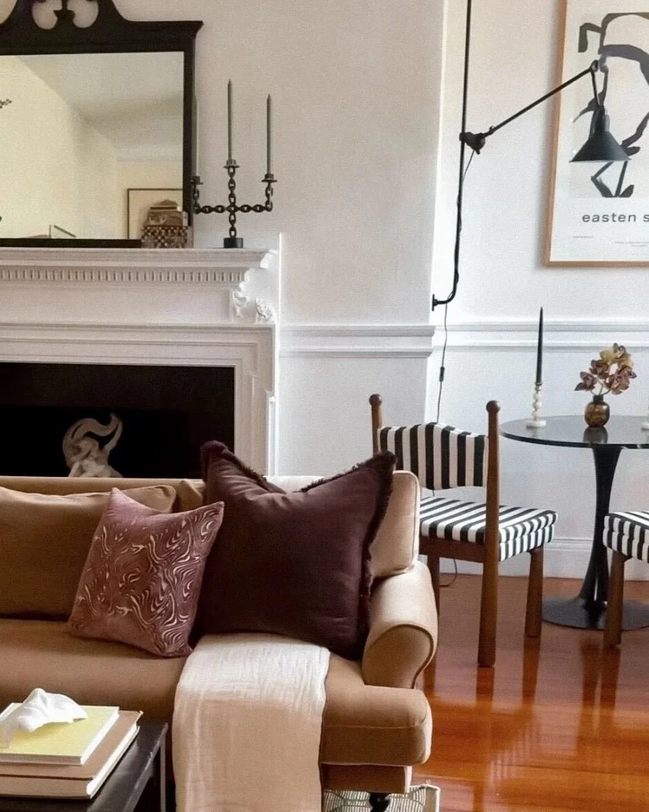 Living room with a beige sofa, decorative pillows, a black side table with books, a fireplace with a white mantel, and a wall-mounted black adjustable lamp. There is a round black table with a vase of flowers, striped chairs, and a framed abstract art piece on the wall.