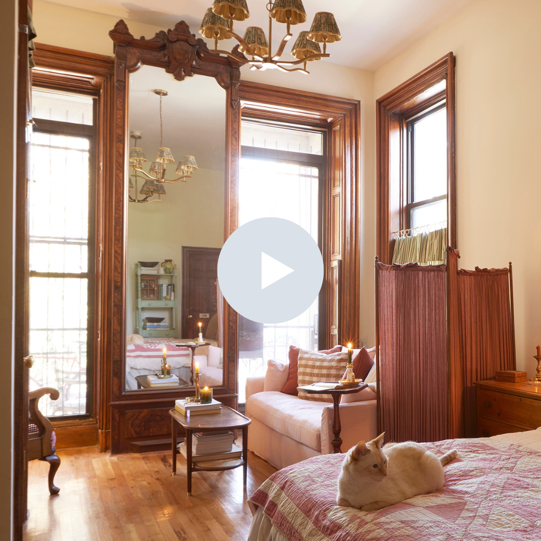 A cozy bedroom with wooden trim, large windows, a bed with a quilt, a cat resting on the bed, a sofa with pillows, a small side table with books, and a vintage mirror reflecting part of the room.