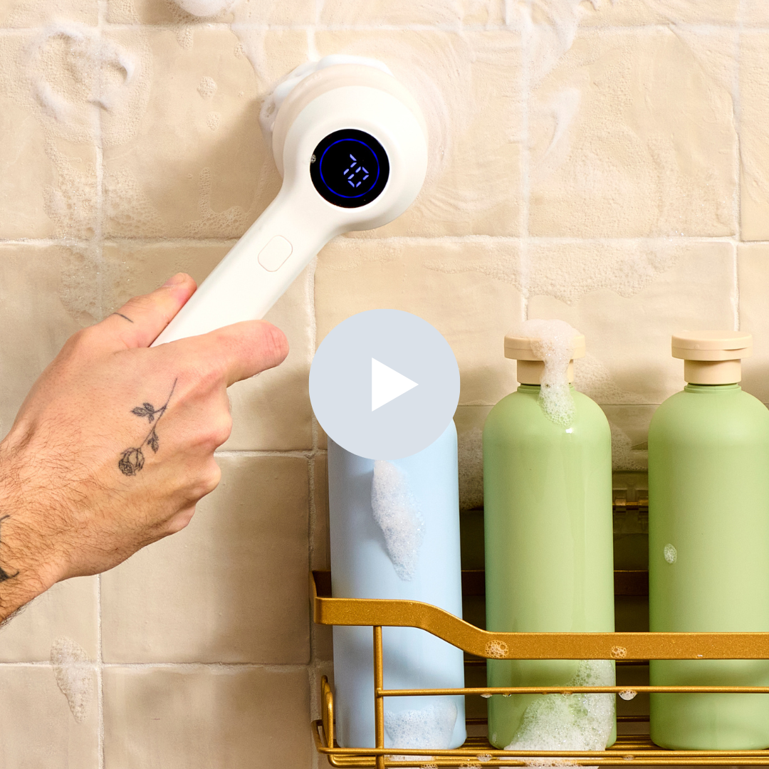 A person using a white electric handheld scrubber to clean soap from a shower wall with three bottles of soap or shampoo in the shower.