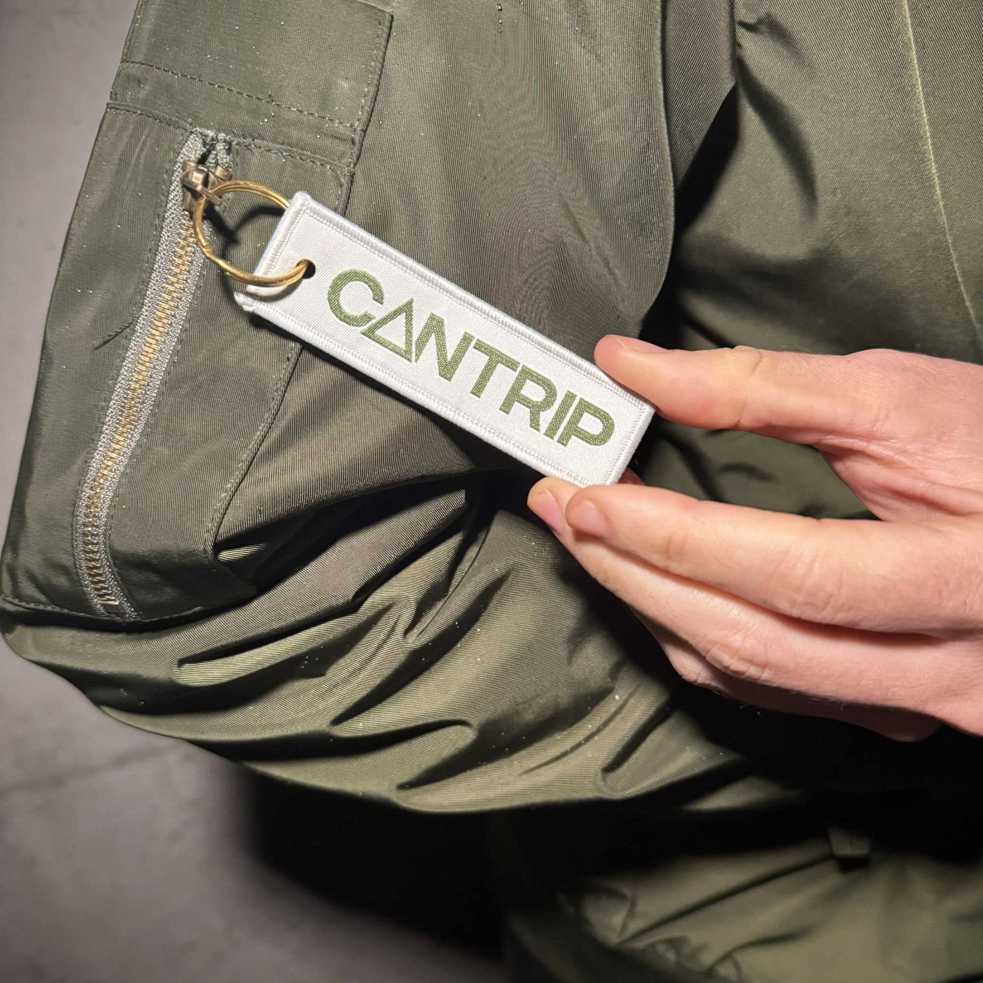 Close-up of a dark green jacket with a zipper pocket. A person's hand is holding a keychain with a white tag that has green text reading 'CANTRIP' attached to the backpack.
