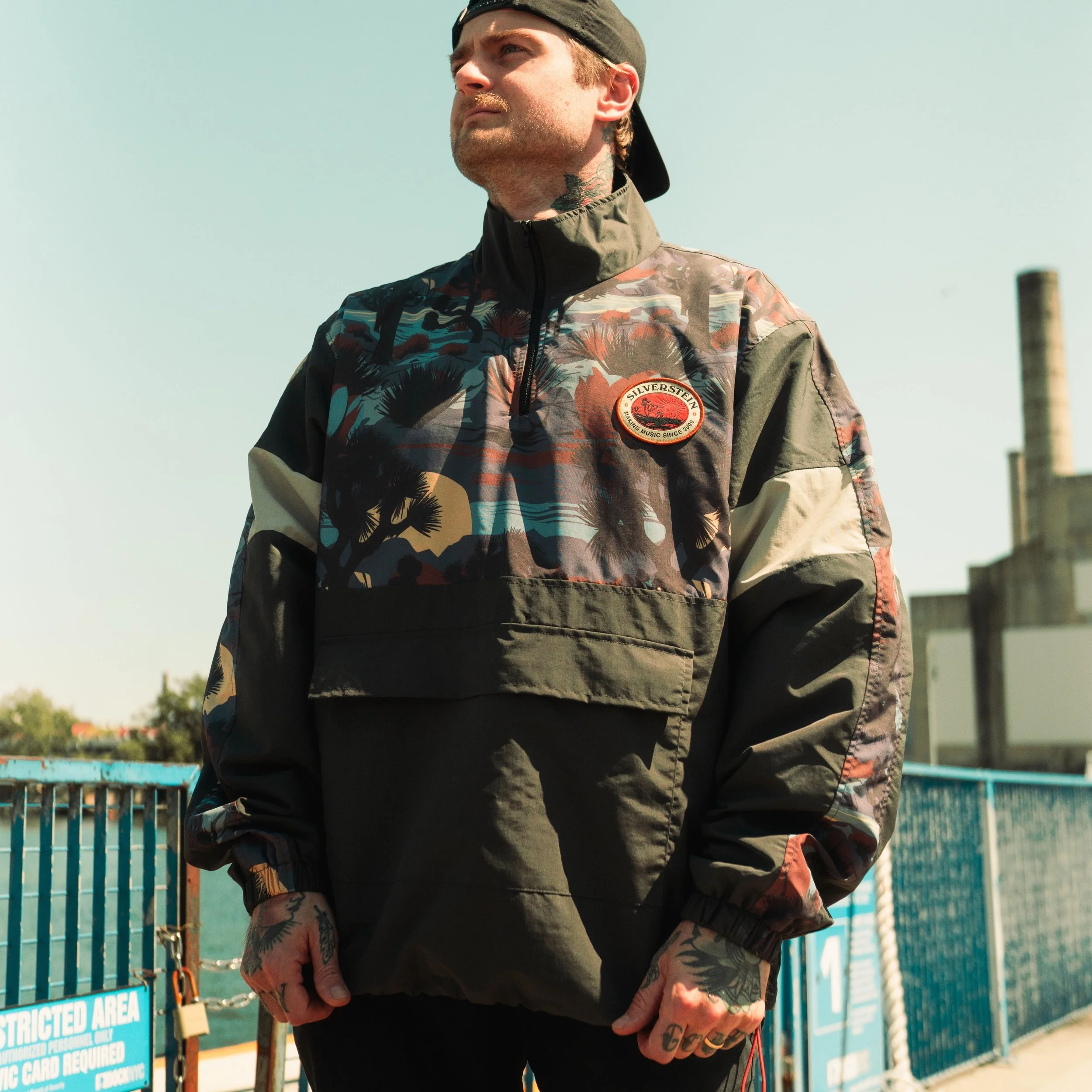 A man with tattoos on his hands and neck, wearing a camouflage jacket with a patch that reads "Silverstein" and a black cap, standing outdoors near a blue fence with a sign that says "Restricted Area."