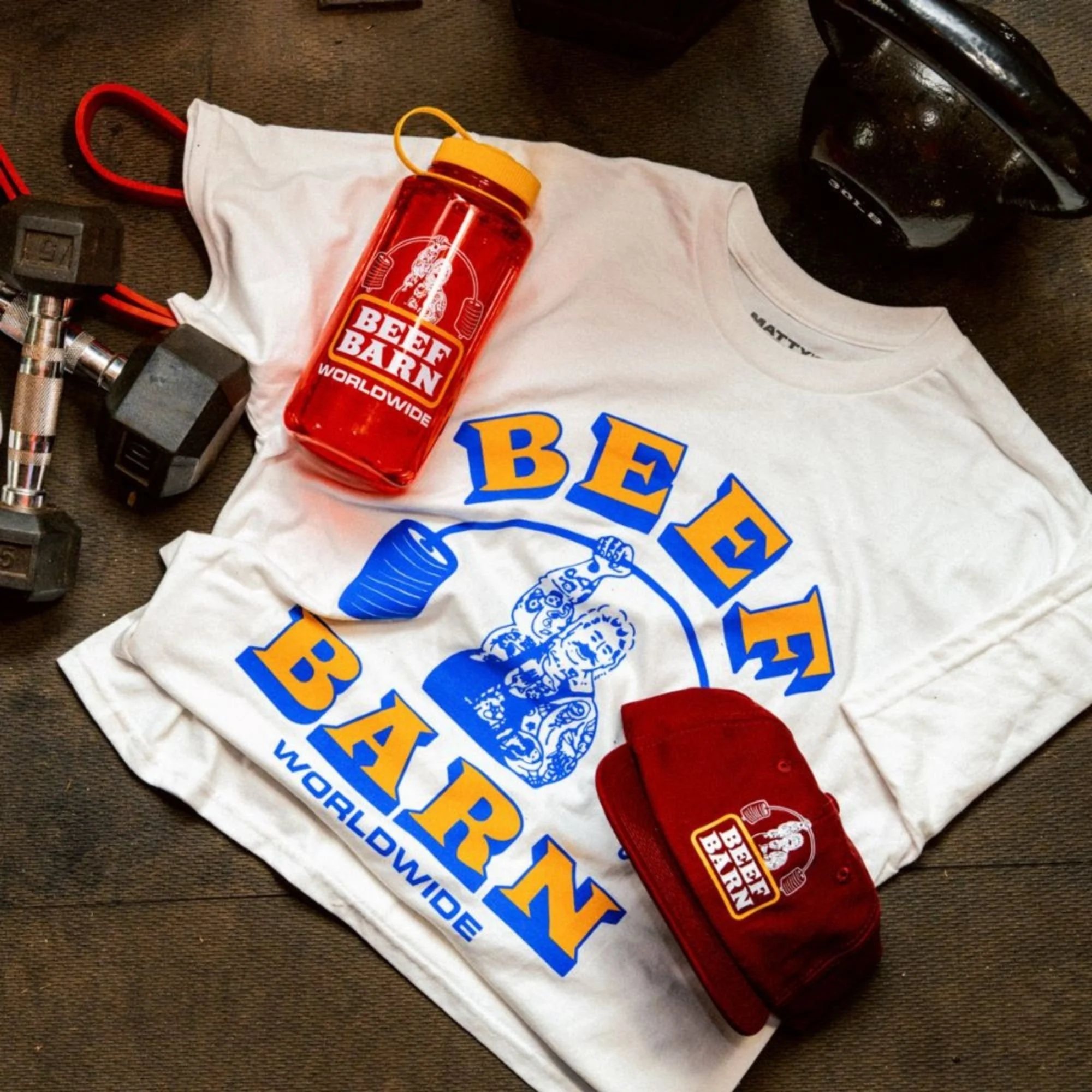 A white T-shirt with a logo, a red drink bottle labeled 'Beef Barn Worldwide', a pair of black dumbbells, a black kettlebell, and a red grip strengthener are laid out on a dark surface.