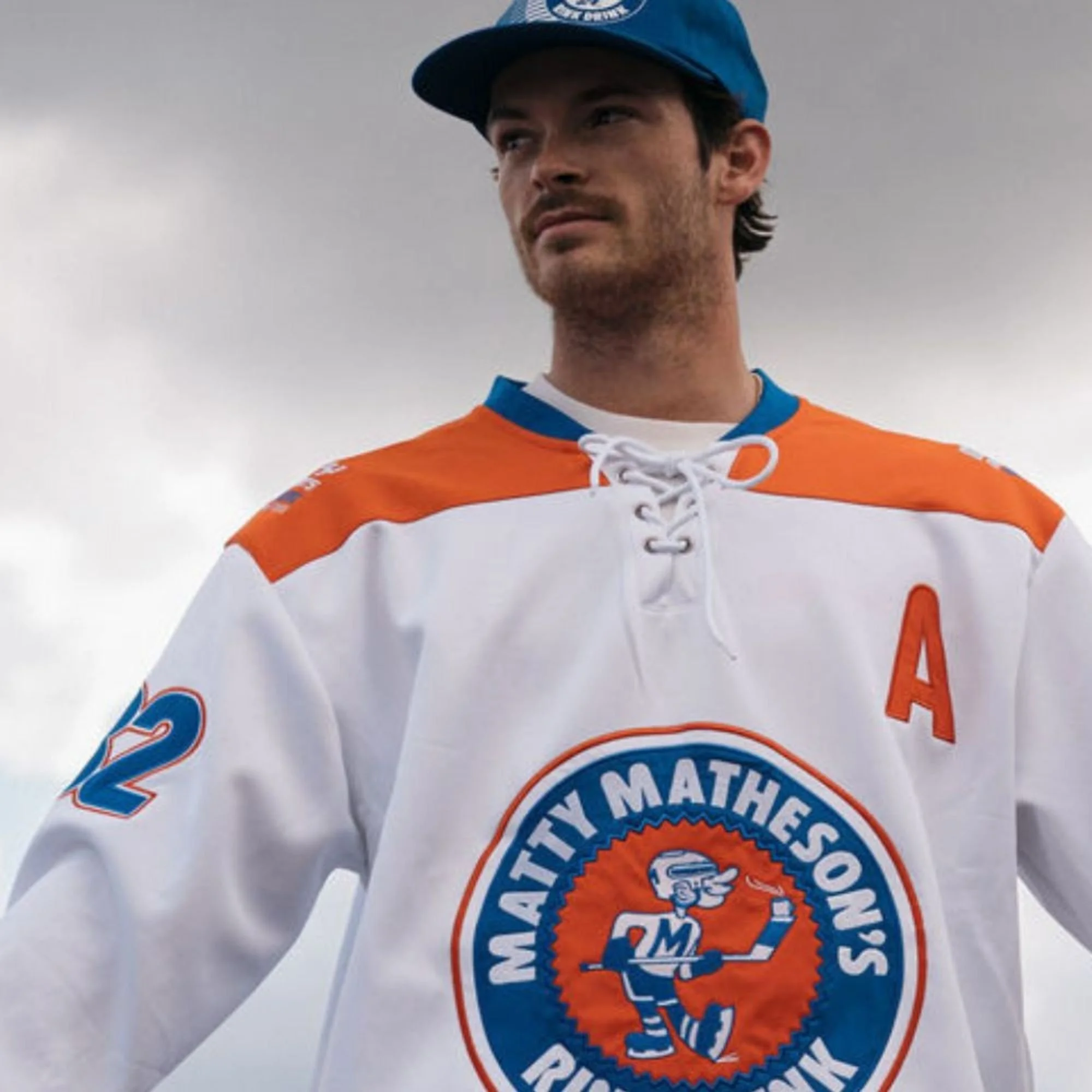 Close-up of a person wearing a custom orange and white hockey jersey with a blue knit beanie, standing against a cloudy sky.
