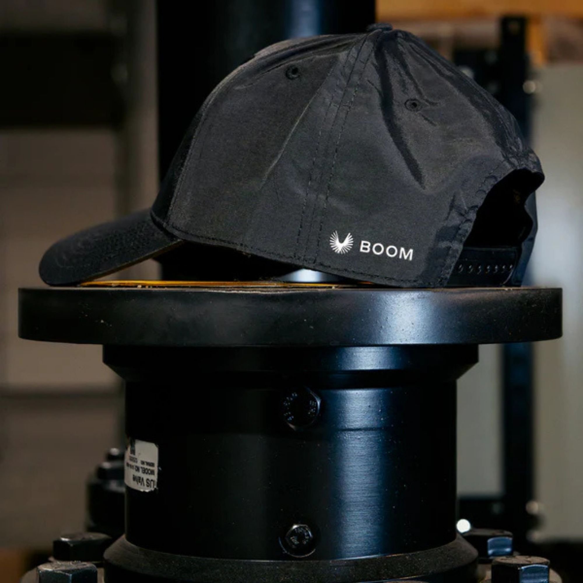 Black baseball cap with curved brim and ventilation eyelets, placed on display stand. The cap features a white logo with the text 'BOOM' and a stylized image resembling a blooming flower or explosion.