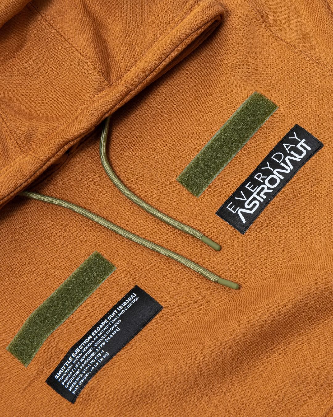 Close-up of an orange Everyday Astronaut hoodie with custom patches, green drawstrings, and Velcro patches.