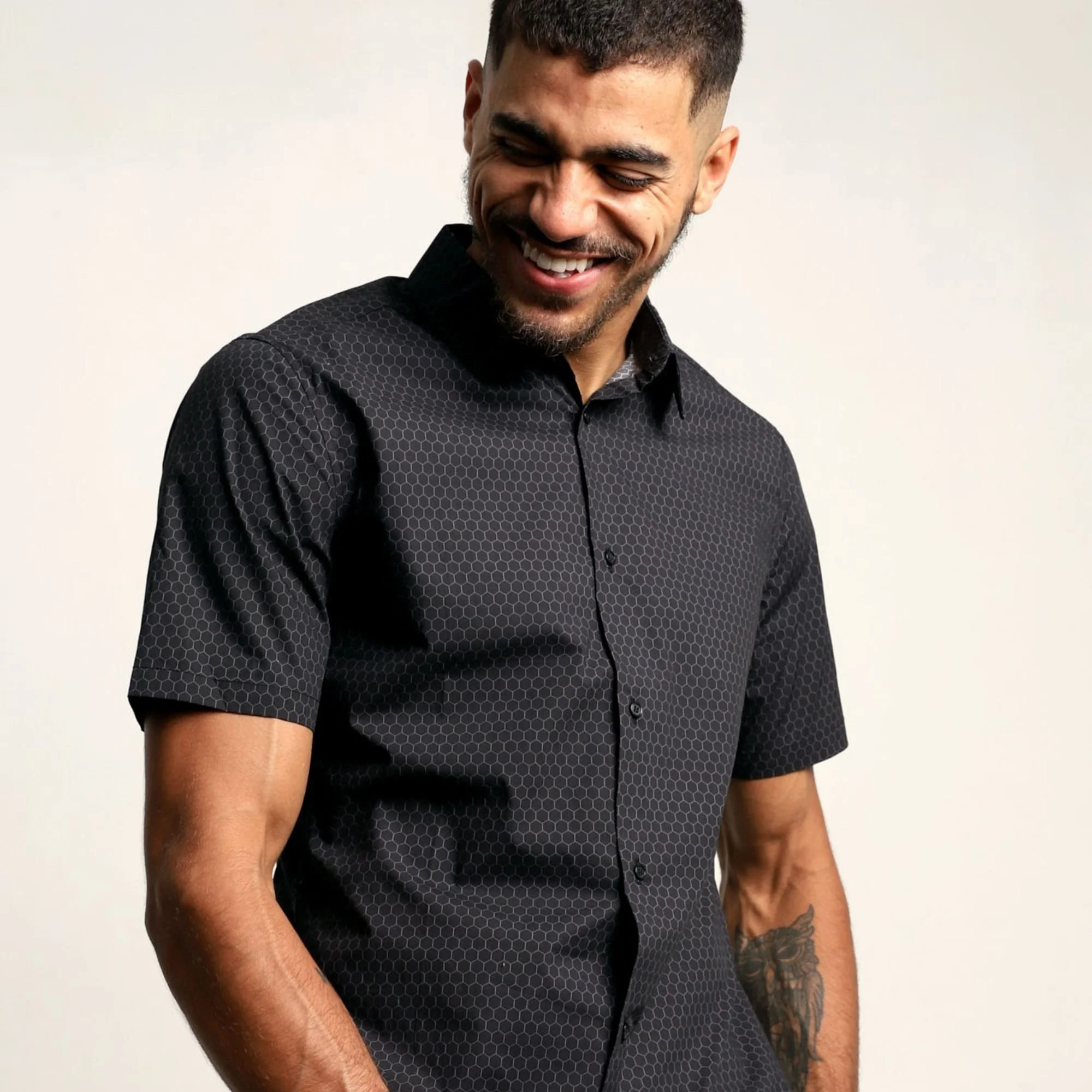 A man with short dark hair and a beard smiling and looking down, wearing a black shirt with a custom honeycomb pattern, against a plain off-white background.