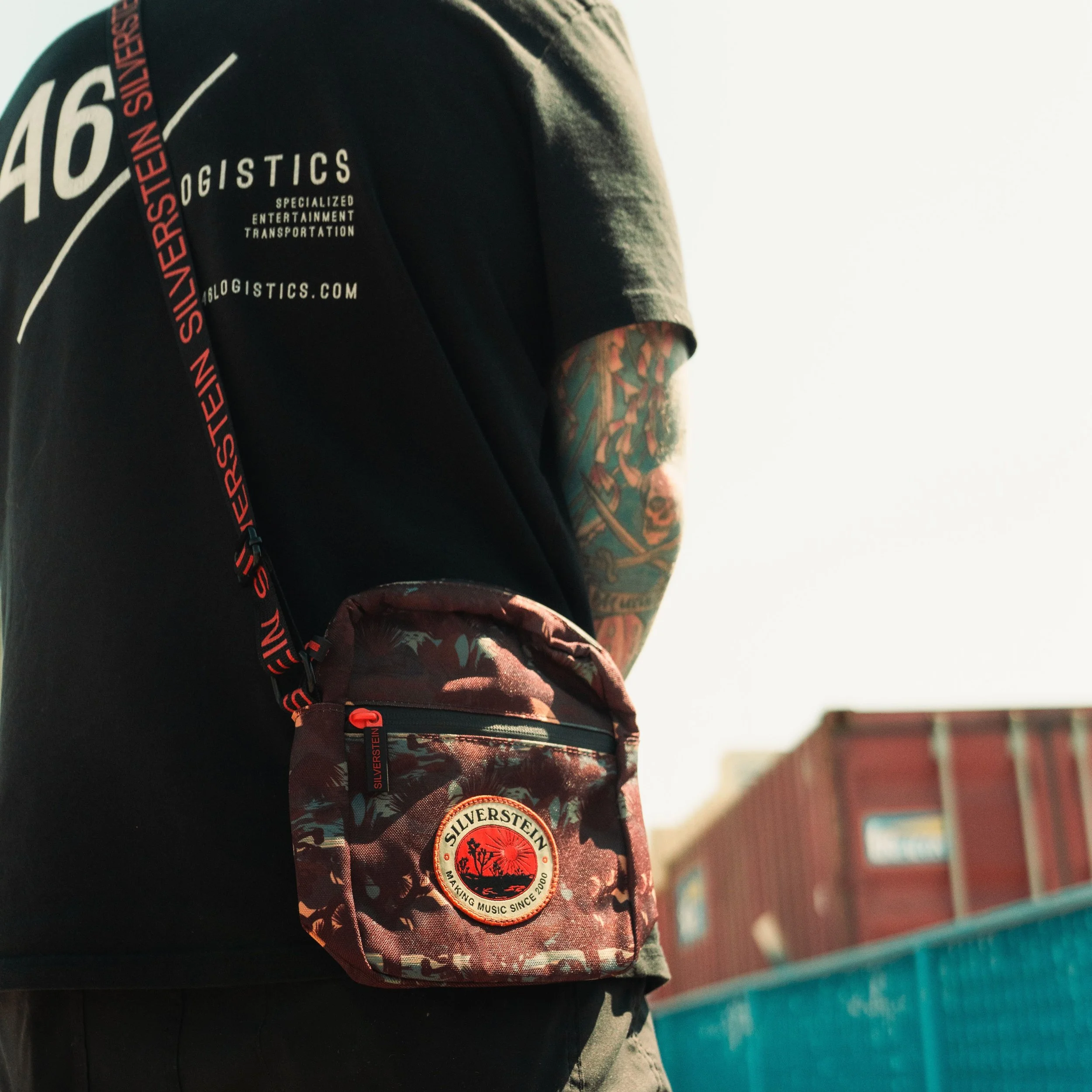 Close-up of a person carrying a camouflage side bag with patches and a black t-shirt with text. The person has tattoos on their arm and is standing in front of a colorful background with buildings.