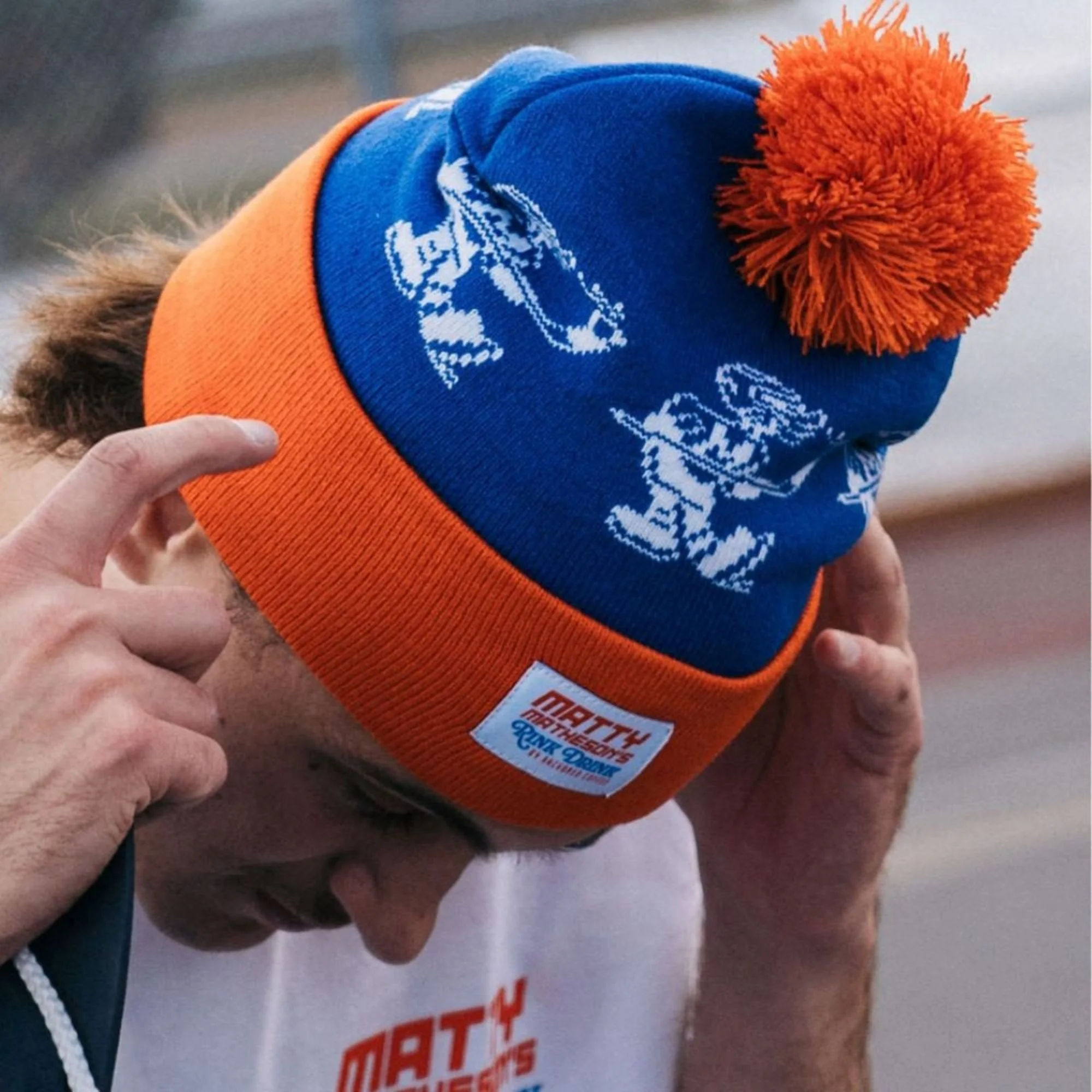 Close-up of a person holding a blue and orange knit beanie with a pom-pom on top, featuring a white logo of a running figure.