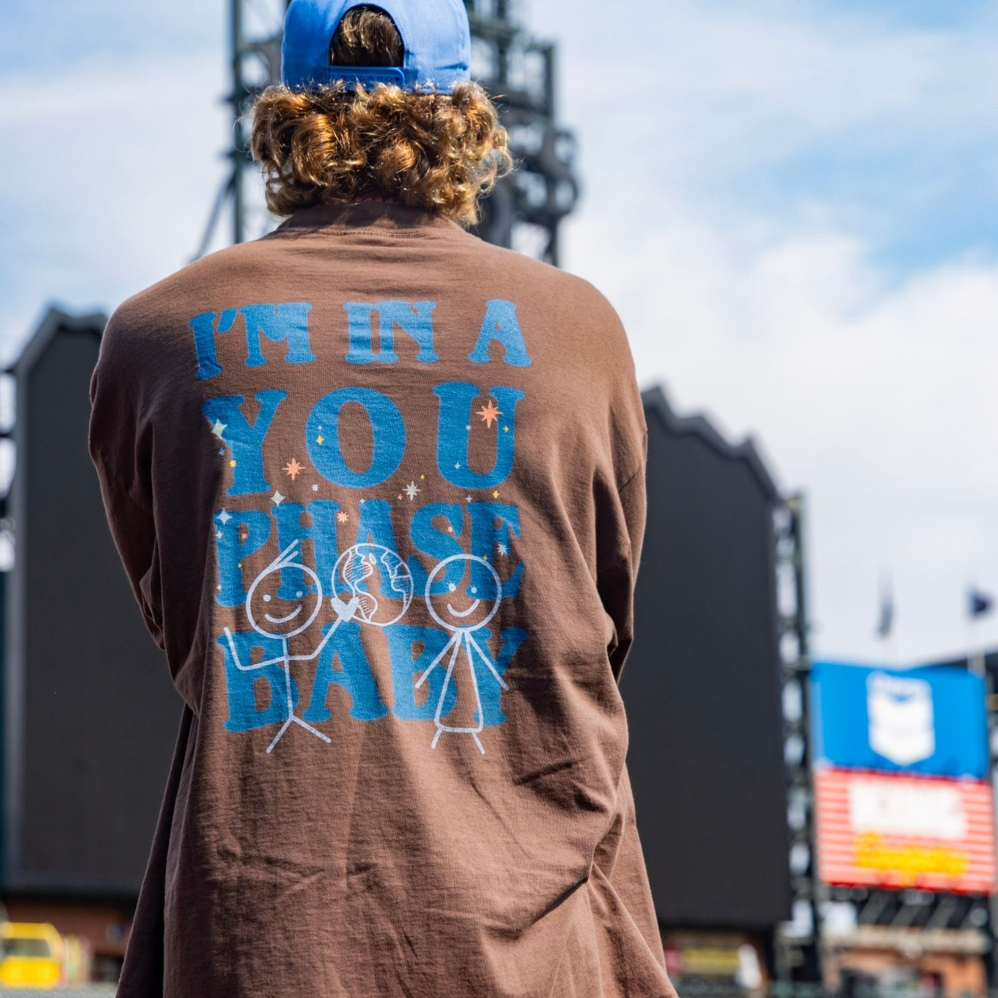 Person with curly hair wearing a brown sweatshirt with blue and white text that says 'I'm in a You Phase" on the back.