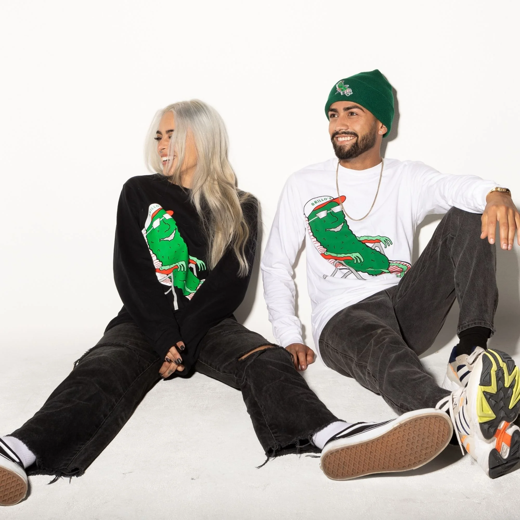 Two people sitting on the floor against a white background, both wearing white shirts with a green graphic. The woman has long, platinum blonde hair and is laughing, wearing black pants. The man has a beard, wearing a green beanie and gray pants, smi