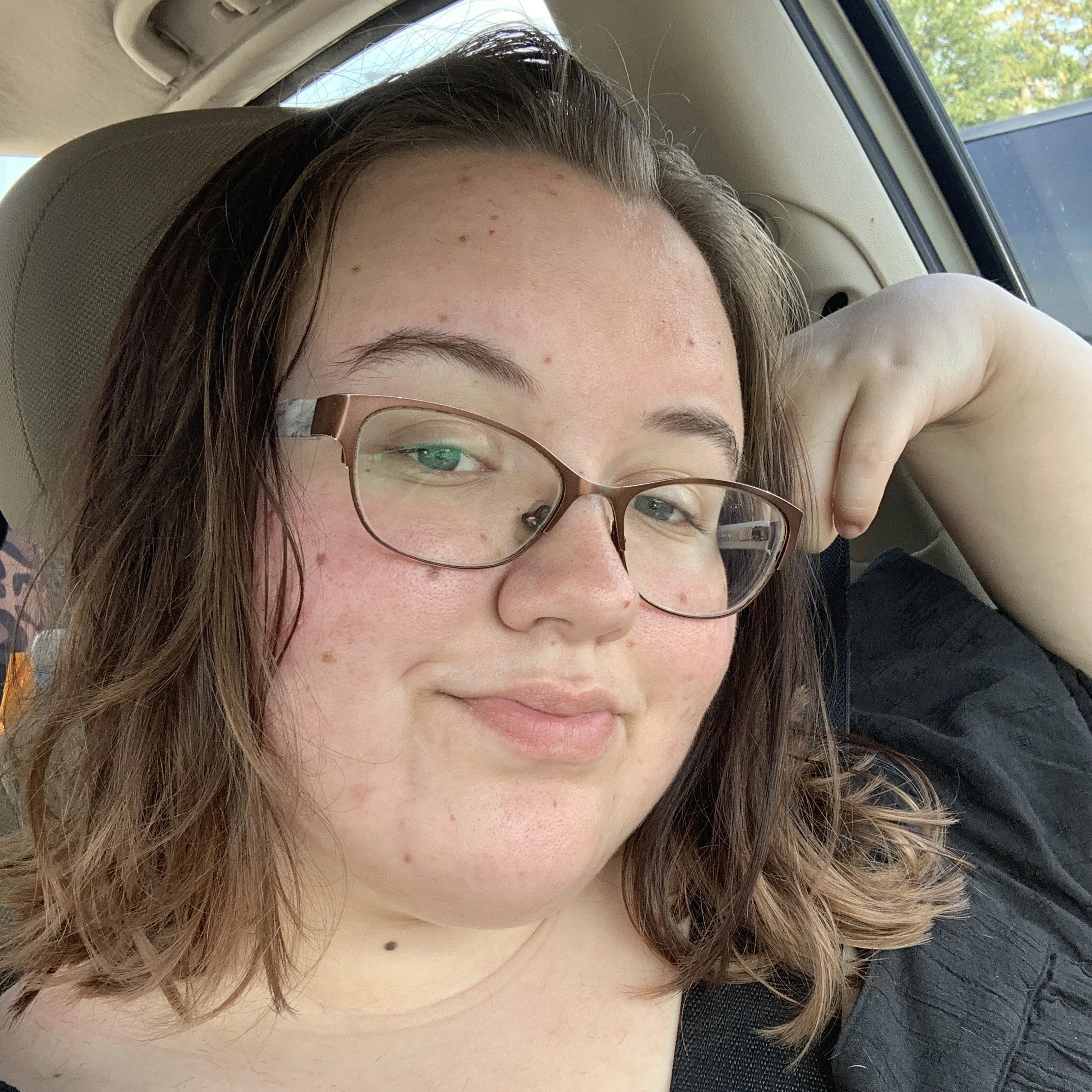 Close-up of a young woman's face with glasses, brown hair, and freckles, lying in a car seat with her arm resting behind her head.