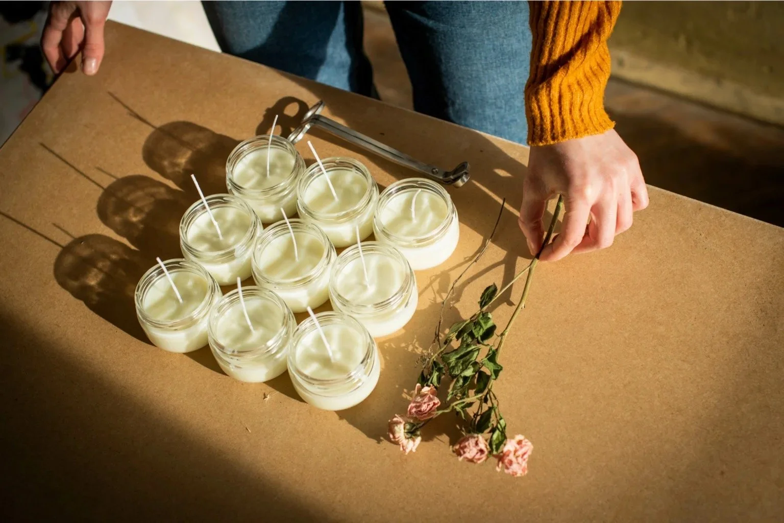 A person placing pink roses on a table with nine lit white candles in glass jars, a lighter, and tongs.