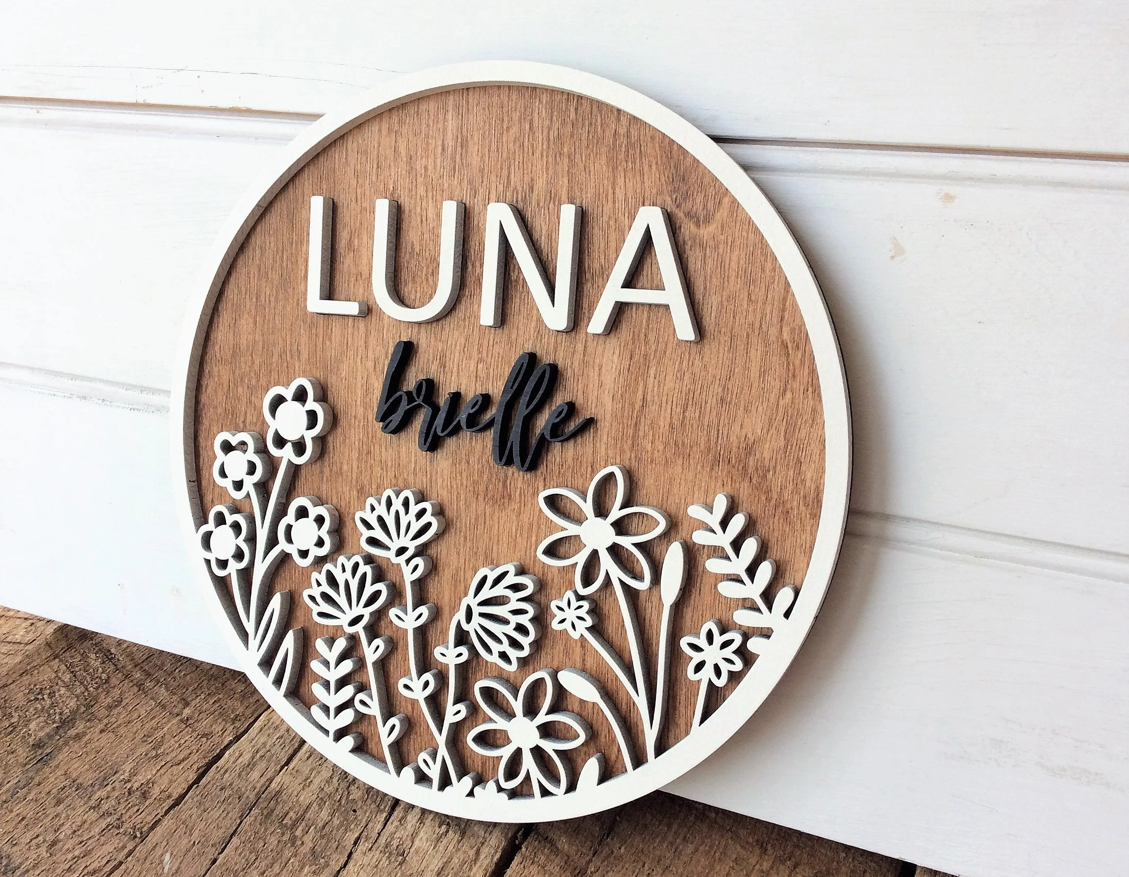 Decorative round wooden wall hanging with the name "LUNA" in white 3D letters and floral cutouts on the bottom.