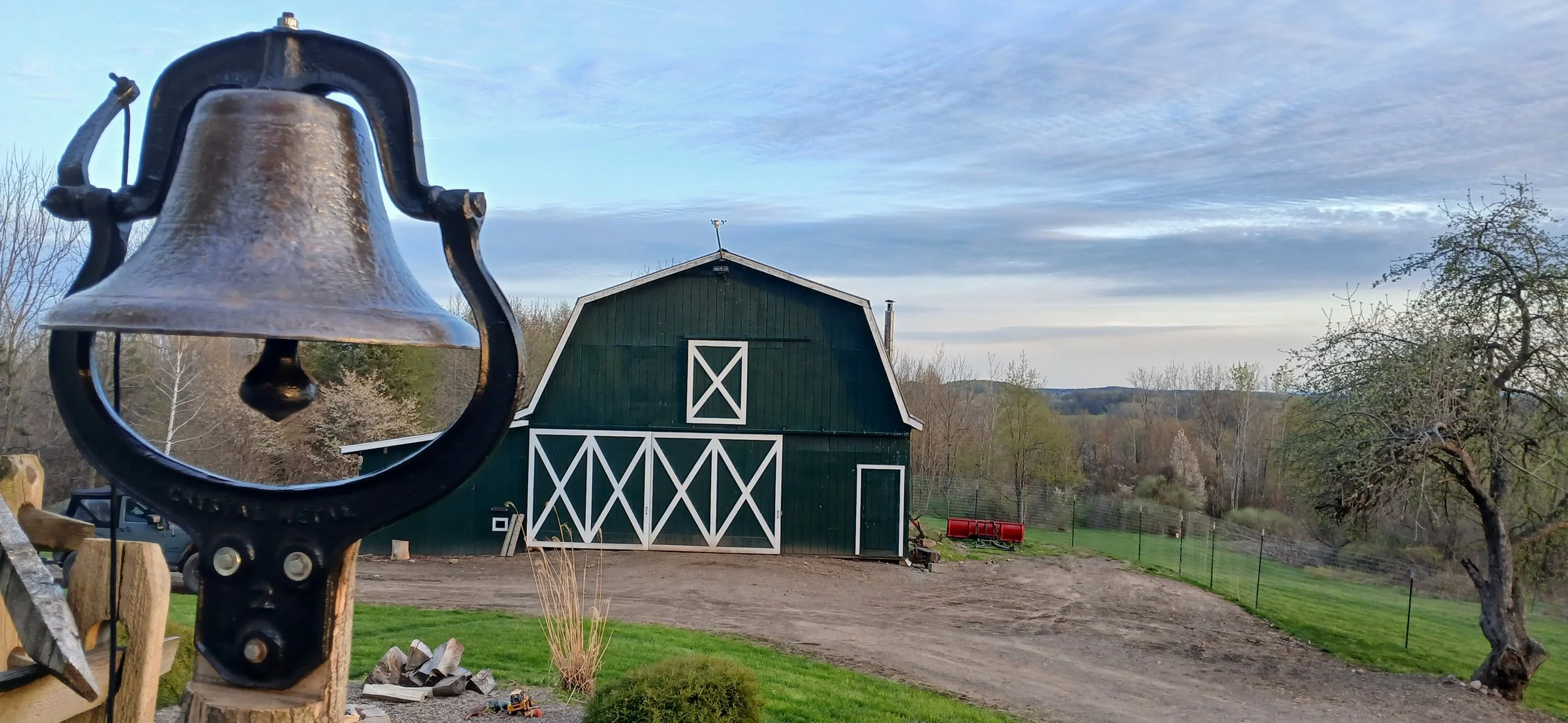 A rural farm scene with a large green barn with white trim, a stone driveway, and trees in the background. A black metal bell is in the foreground, mounted on a wooden post.