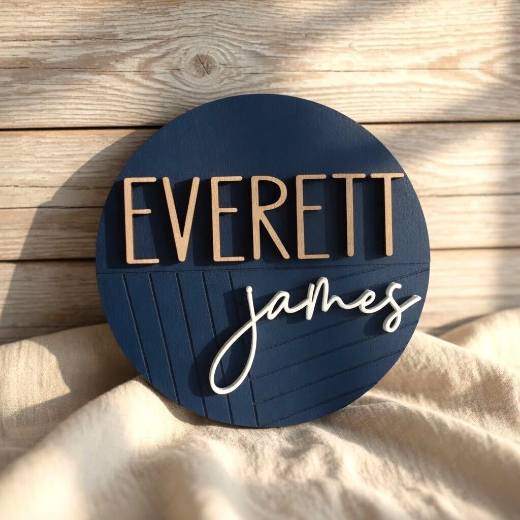 A round dark blue sign with the words "Everett James" in large wooden and white cursive letters, resting against a light wooden background and a cream-colored fabric.