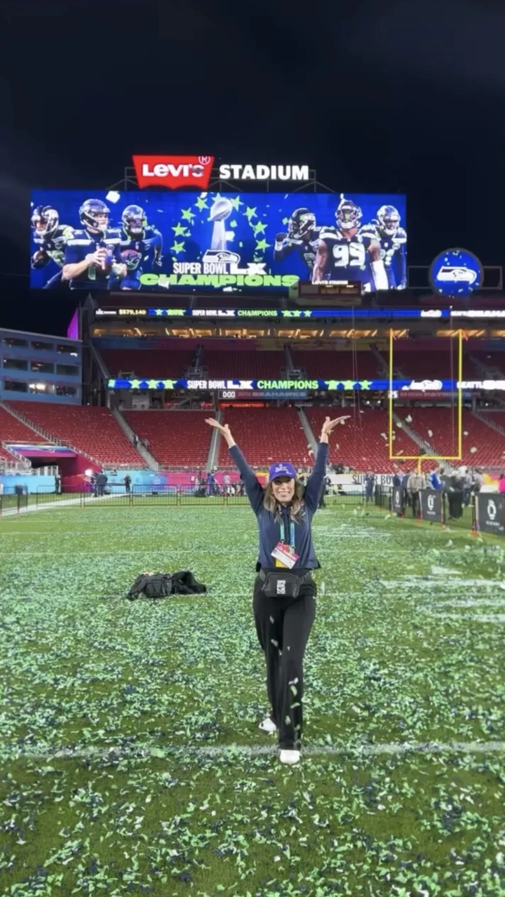 A woman celebrating on a football field covered in confetti after the Super Bowl victory, with a large screen showing football players and the words "Super Bowl Champions" in the background.