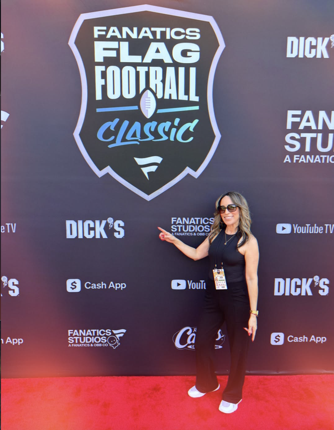Woman in black outfit with sunglasses pointing to a large banner for Fanatics Flag Football Classic event, featuring various sponsor logos.