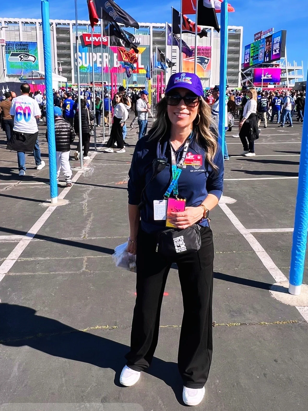 A woman smiling wearing a purple cap, sunglasses, and a dark blue shirt with a Super Bowl badge. She is standing outside in a parking lot with people and flags in the background, celebrating a Super Bowl event at a stadium.