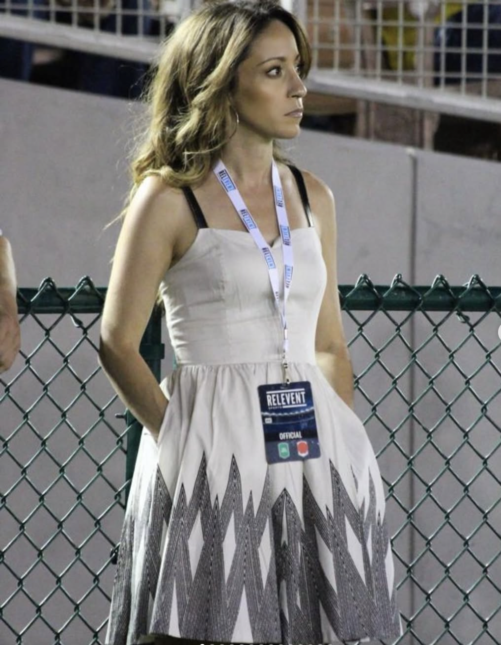 A woman with light brown wavy hair standing outdoors, wearing a beige dress with black straps and a patterned bottom, a lanyard with an event badge, and leaning against a chain-link fence.