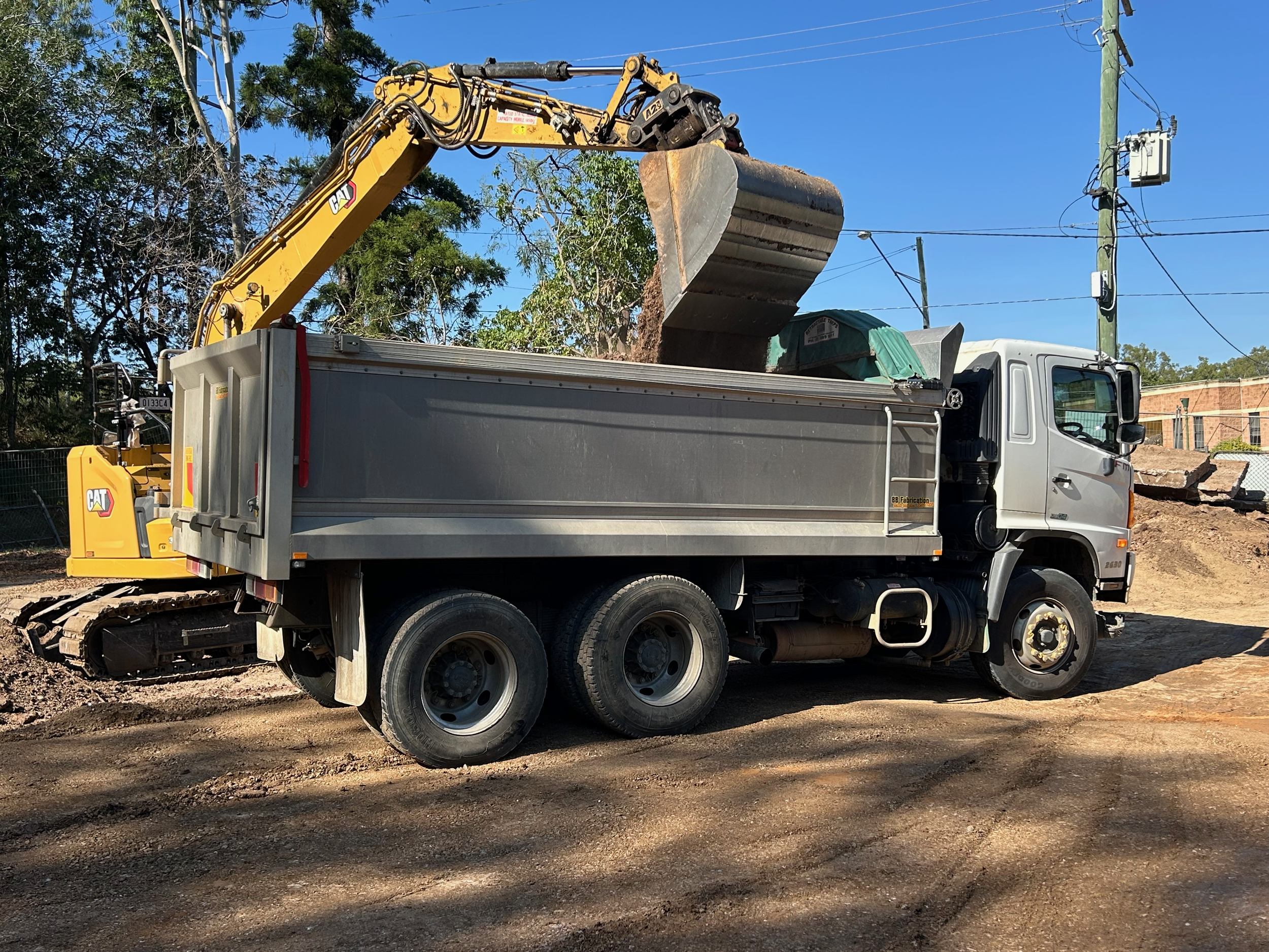 A construction excavator with a yellow arm and black bucket working on a dirt site, mounted on a truck with a gray cab, under a blue sky with trees and utility poles in the background.