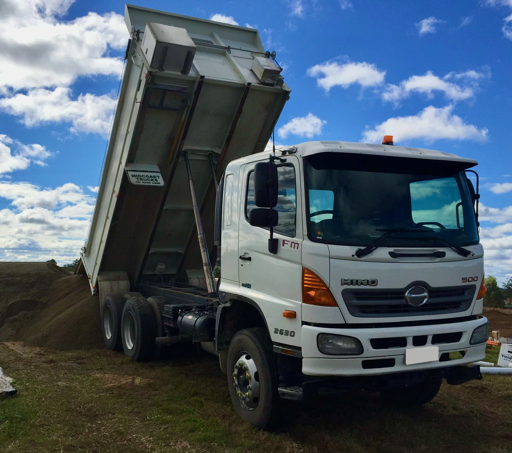 White dump truck with raised bed, dumping dirt or gravel onto the ground during daytime under a partly cloudy sky.