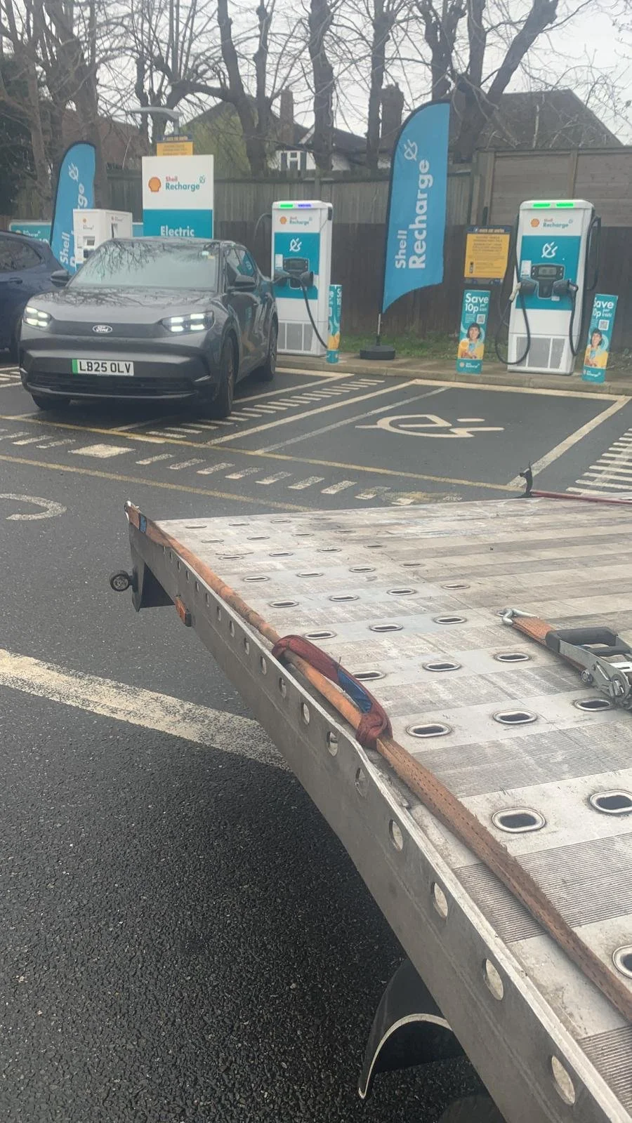 An electric vehicle is plugged into a Shell Recharge charging station in a parking lot with brand flags, and a flatbed truck is in the foreground.