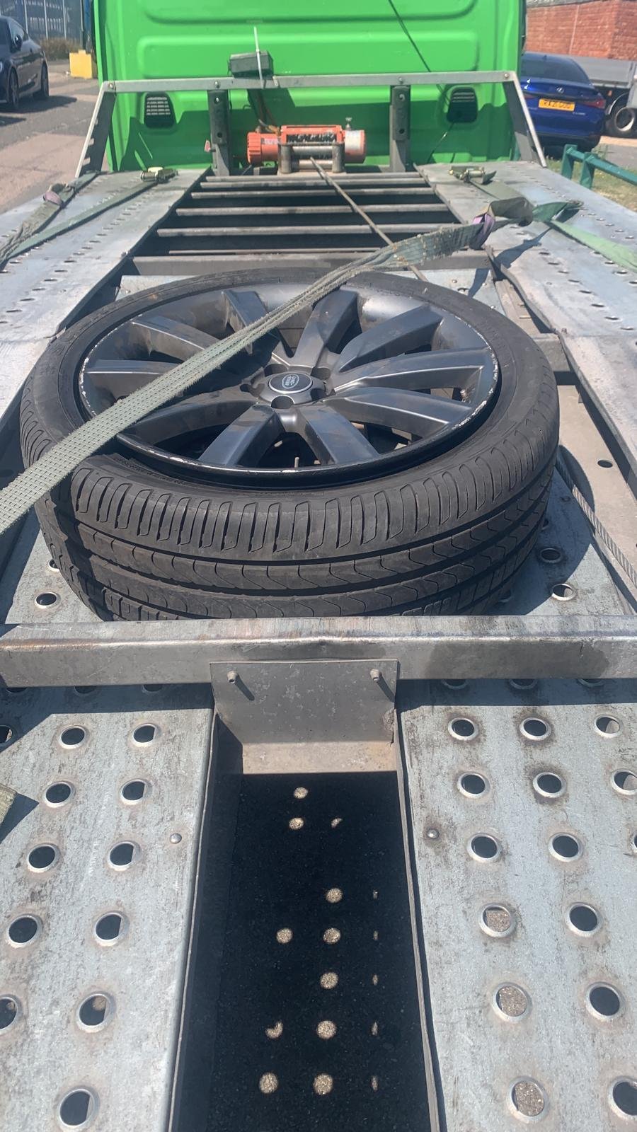 A spare tire with a black alloy wheel on a metal flatbed trailer secured with ratchet straps.