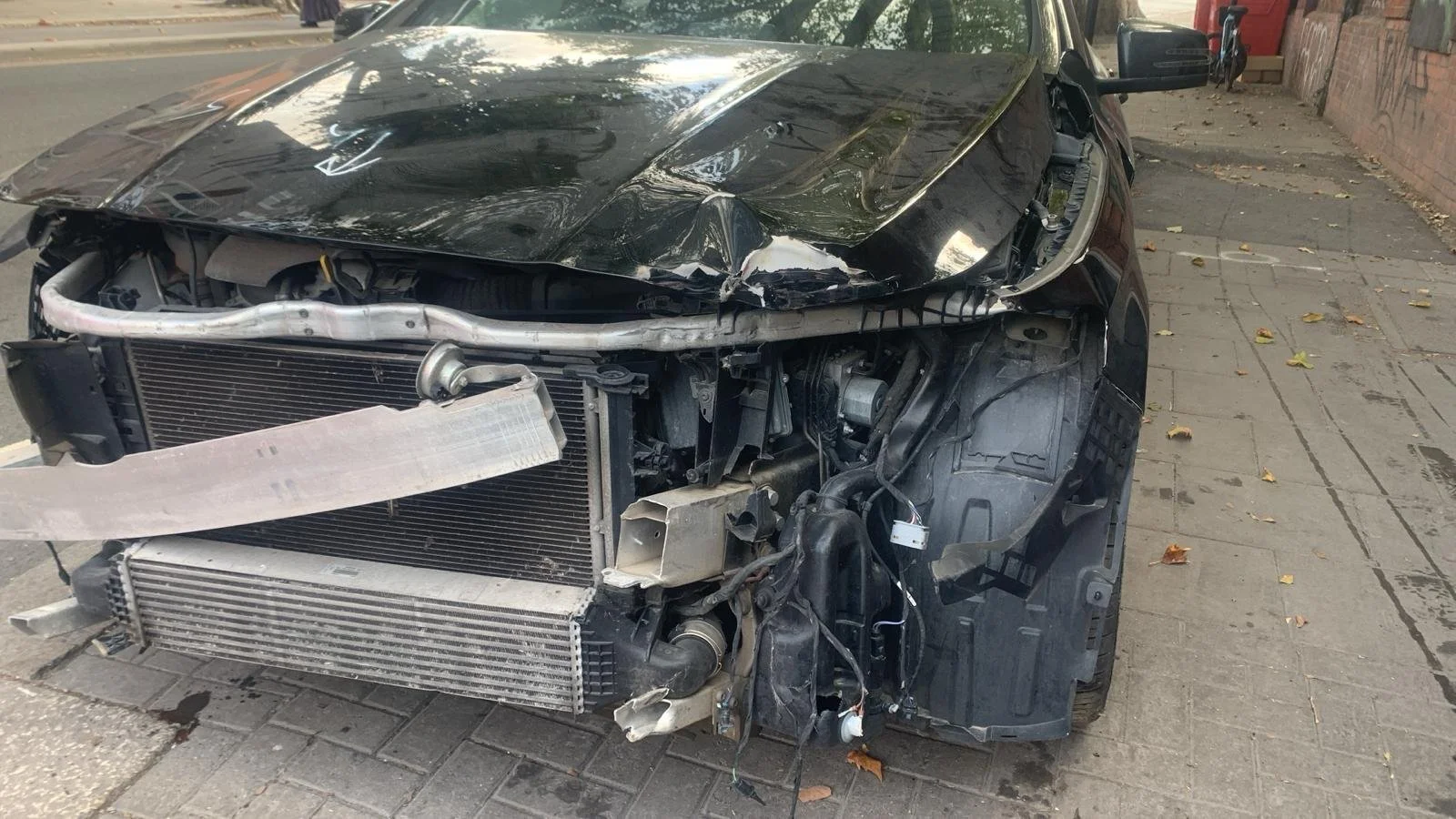 Damaged black car with front end heavily crashed, exposing radiator and internal components, parked on city sidewalk.