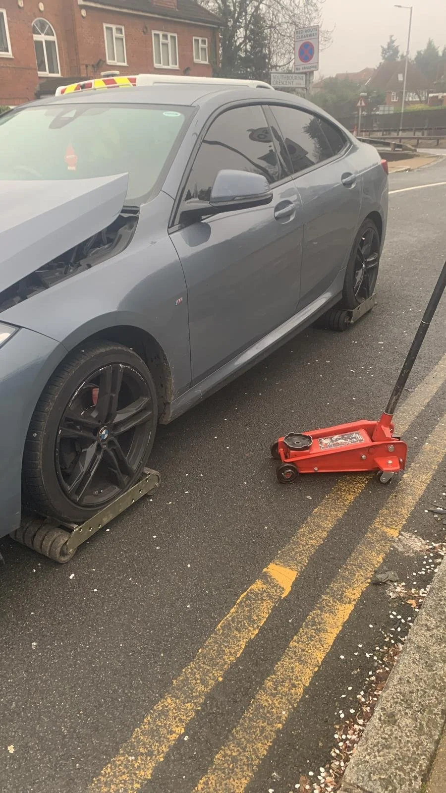 A gray BMW car with the front wheel removed and placed on a roller track, and the rear wheel missing with a wheel clamp attached. A red hydraulic floor jack is positioned nearby on the asphalt street.