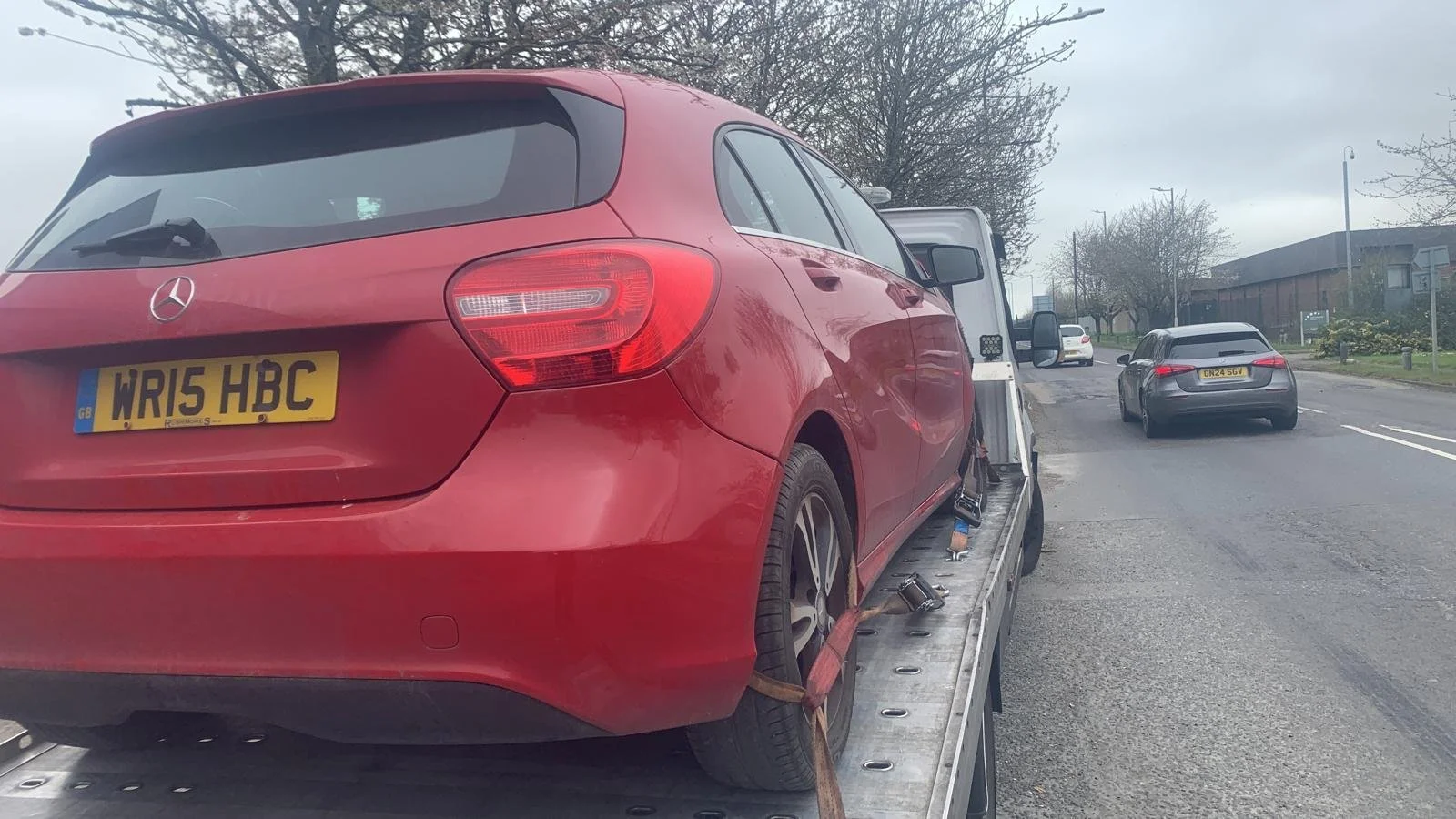Red Mercedes-Benz car loaded onto a flatbed tow truck on the side of a street, with a highway and other vehicles in the background.