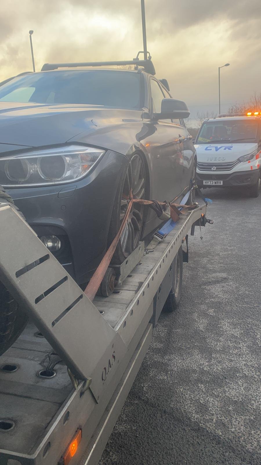 A black car being transported on a flatbed tow truck, secured with straps, with a police vehicle behind it on cloudy weather.