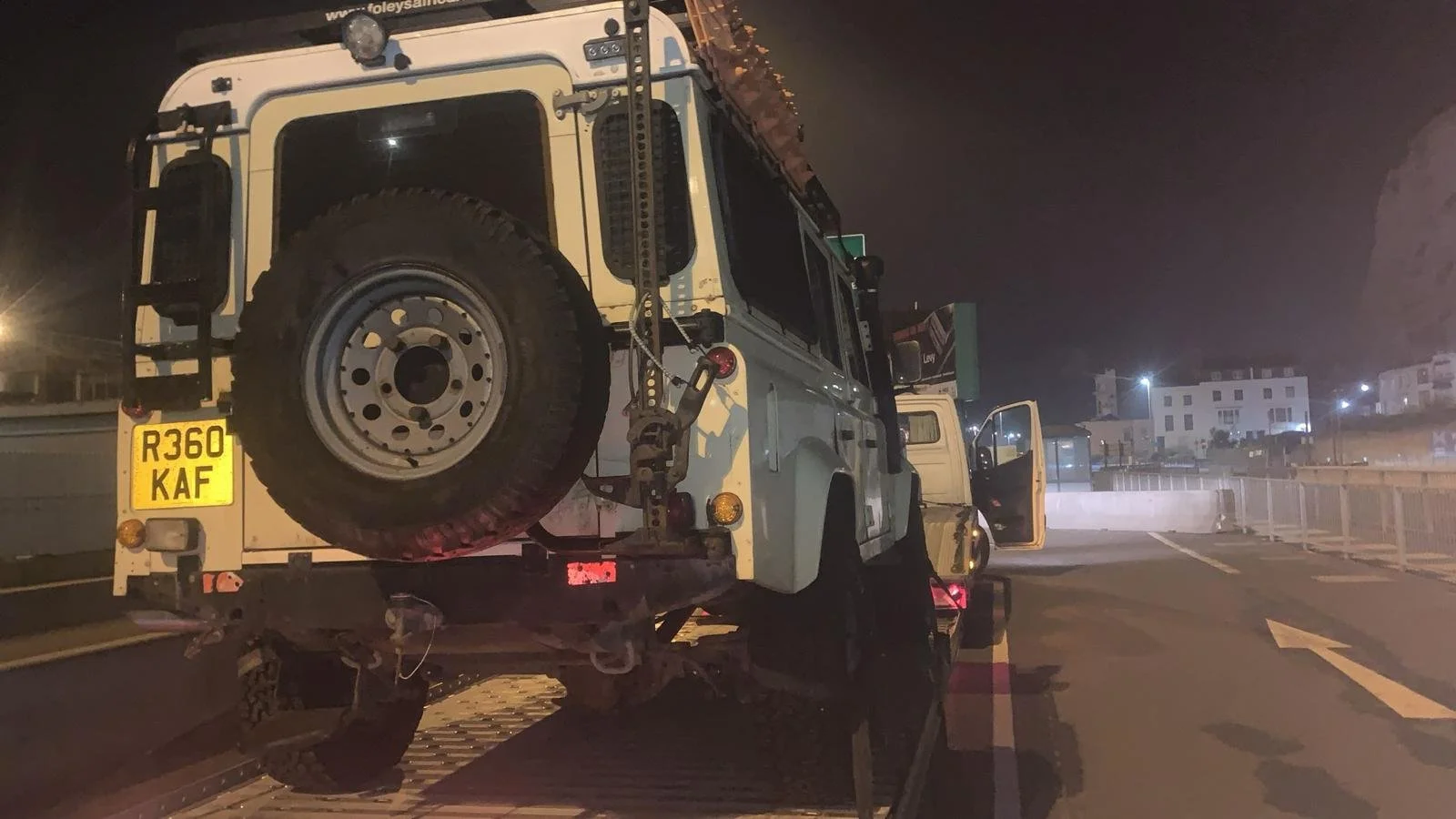 Nighttime scene of a white off-road utility vehicle on a flatbed tow truck, with a spare tire mounted on the back of the vehicle and a hydraulic arm attached. The background shows a dimly lit urban area with buildings and streetlights.