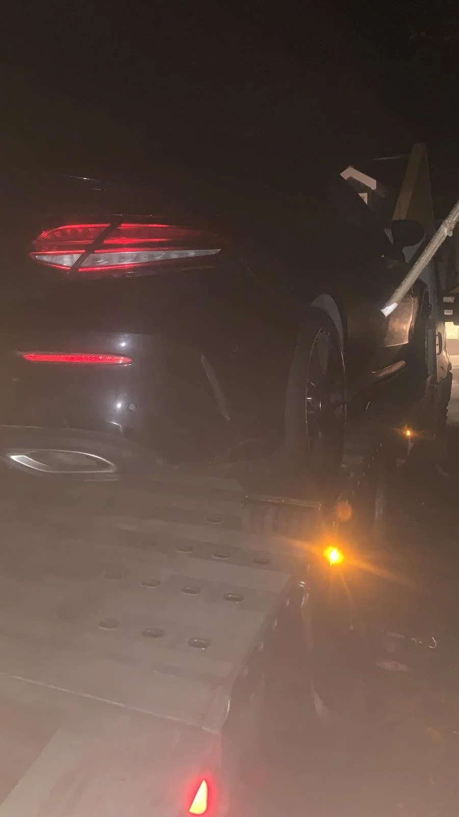 A dark-colored luxury car is being loaded onto a flatbed tow truck at night. The car's taillights are illuminated, and a metal bar or support structure is attached to the car, indicating it is being towed.