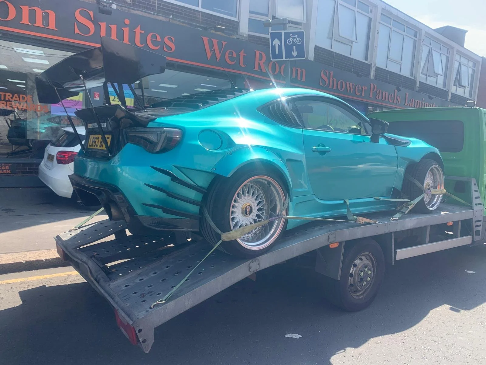 A blue sports car secured with straps on a flatbed tow truck, parked outside a store with signage advertising wet rooms and shower panels.